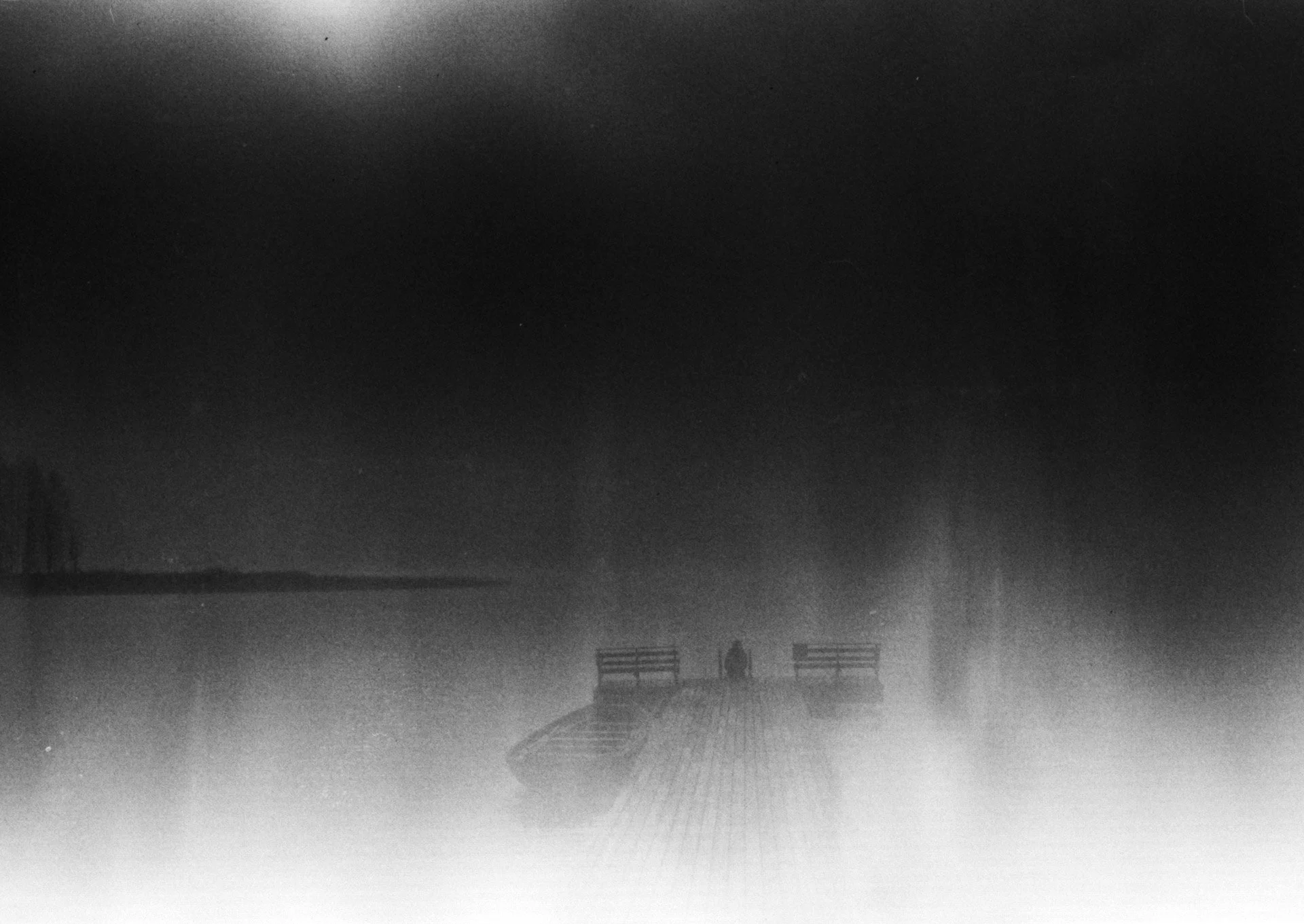 A foggy night scene of a wooden pier extending into a body of water with one person standing at the end. Two benches are located on either side of the pier, and faint structures are visible in the distant background.