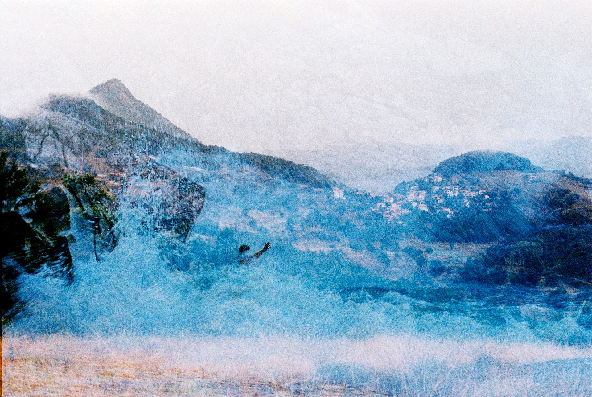 A waterfall flowing down a mountain with a person reaching out in the water, and a town visible in the distance on the hillside.