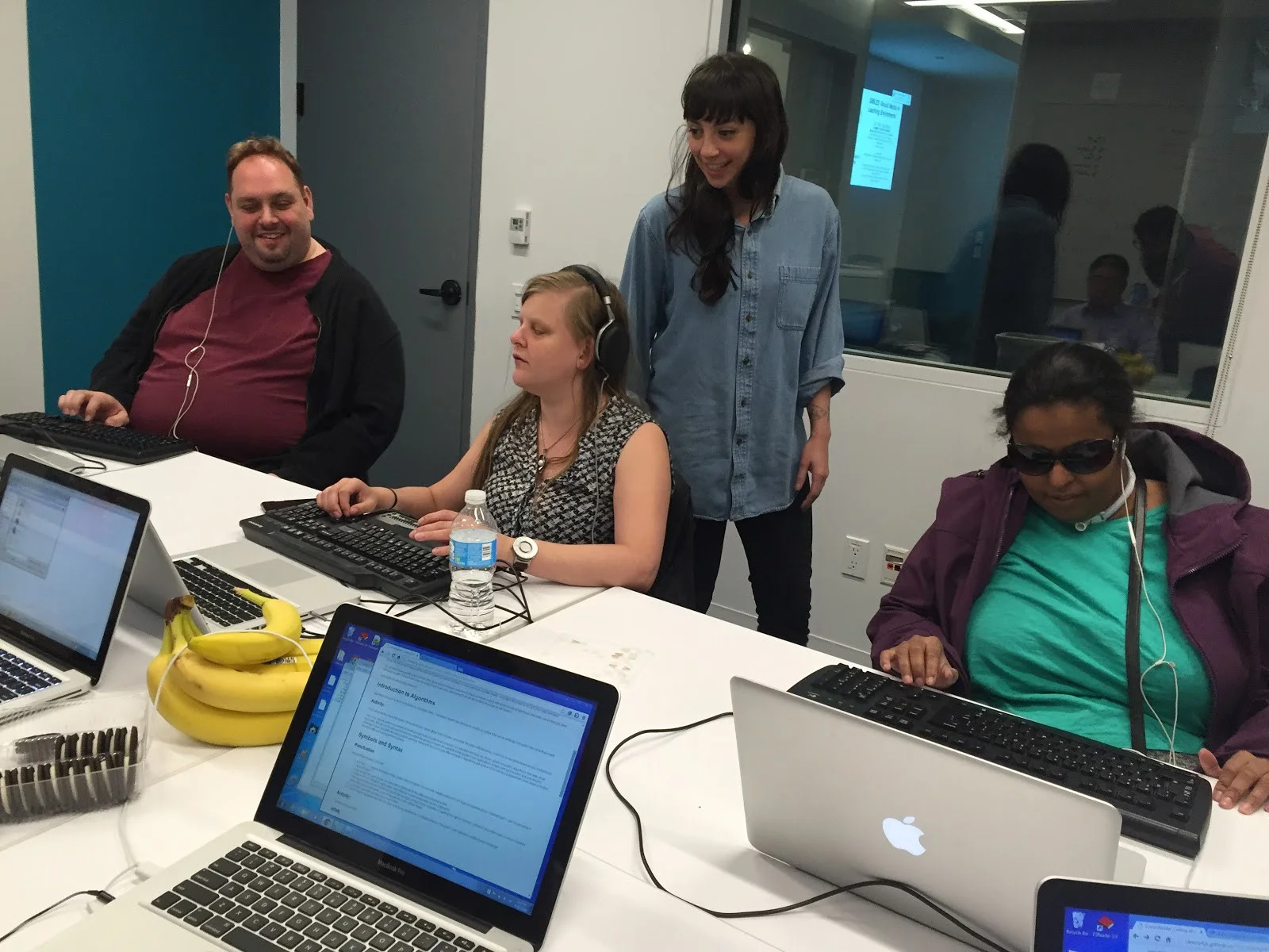 Figure 2. Claire stands behind three workshop participants sitting at a table with laptops in front of them.
