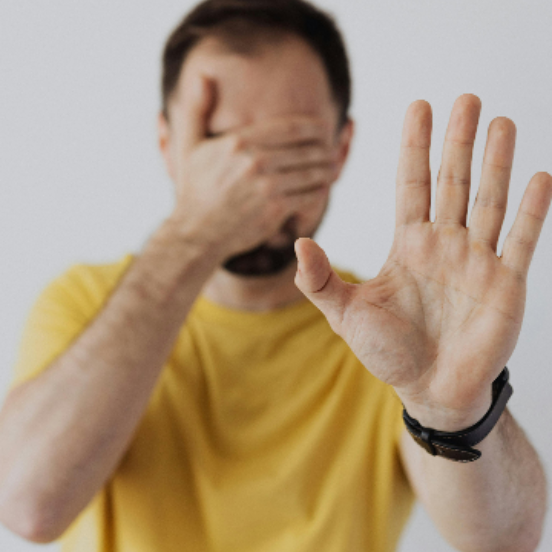 A man in a yellow shirt covering his eyes while with one hand while extending his other arm out with palm facing out to stop something