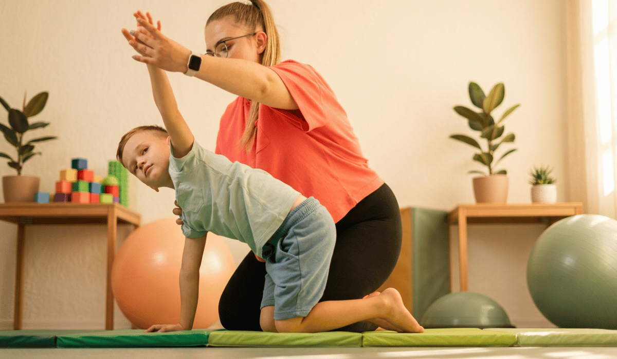 a boy on hands and knees on a gym mat with a therapist helping him.