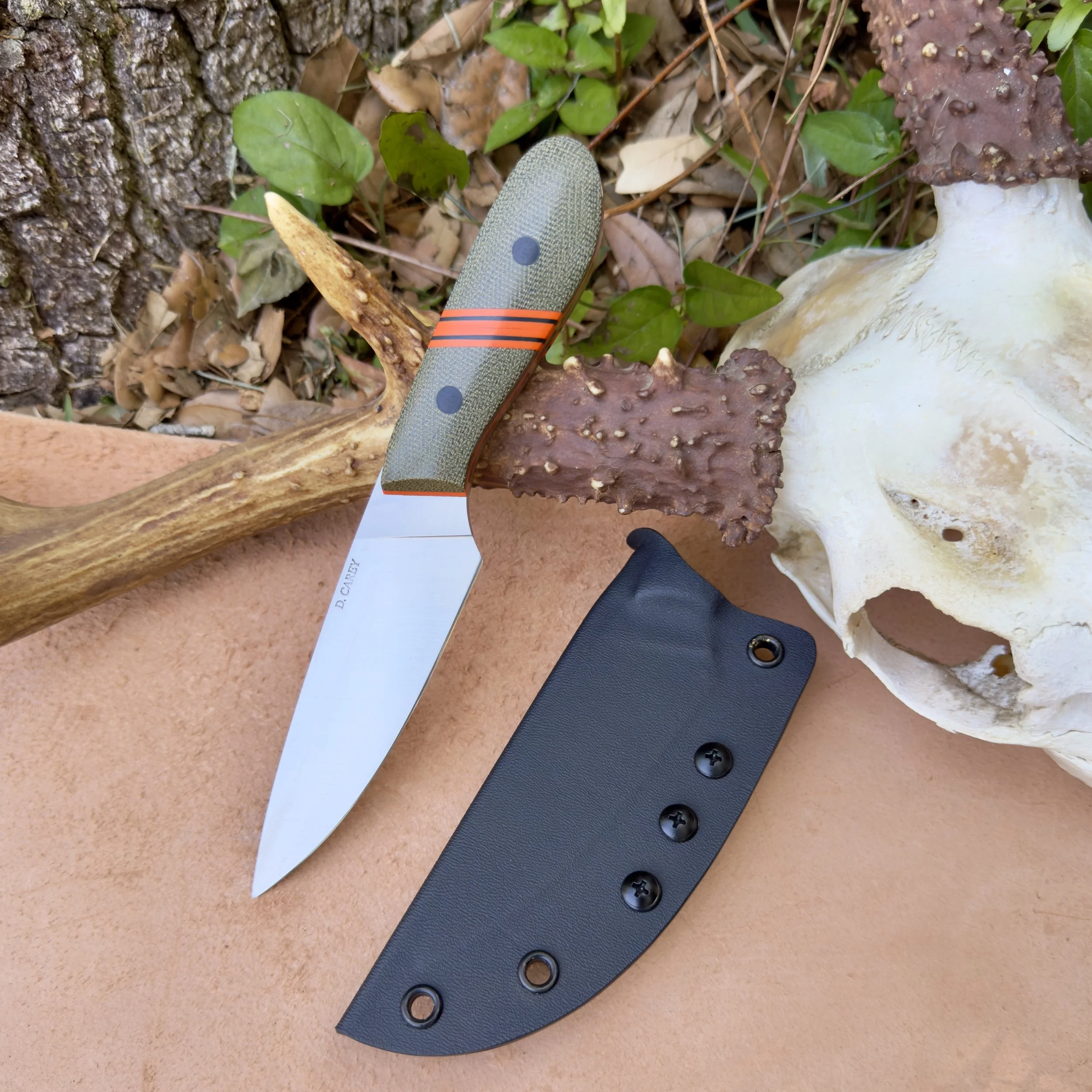 Custom EDC fixed blade knife with CPM-154 stainless steel blade, green micarta handle with orange accents, and black Kydex sheath on outdoor surface