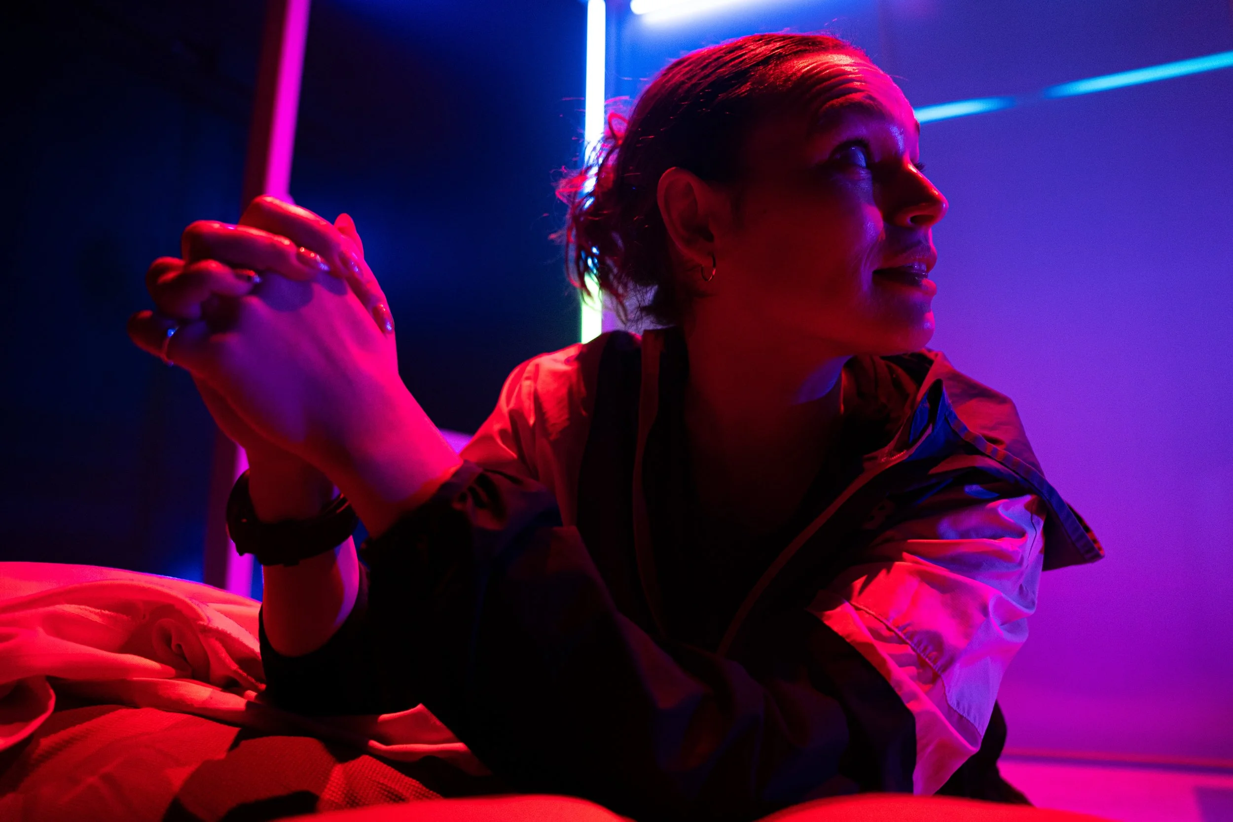 Photograph of a young woman looking over her shoulder while her hands are clasped in prayer. She is illuminated by a vibrant pink light.