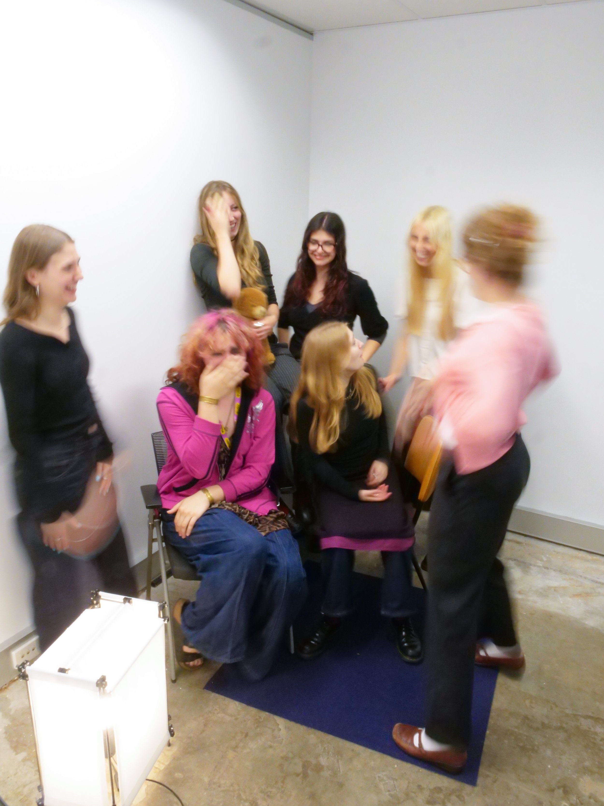 Photo of 7 figures dressed in pink, black, grey and white. 2 are sitting down on chairs and 5 are standing around them. Some parts of the photo are blurry from motion.