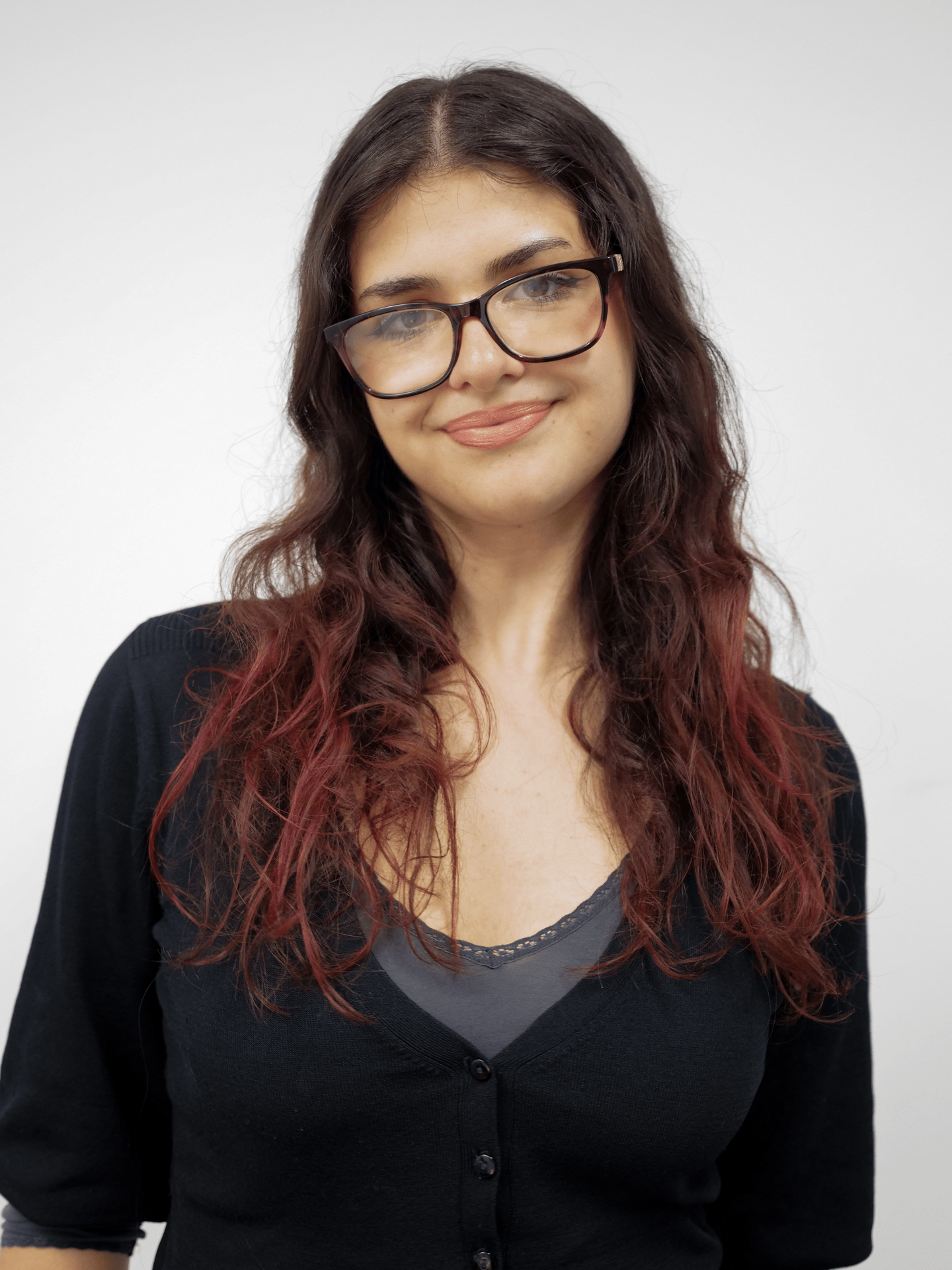 A gif cycling through several portraits of a figure. The figure has long black and dark red wavy hair. They are wearing large black glasses, a grey top with a black cardigan.