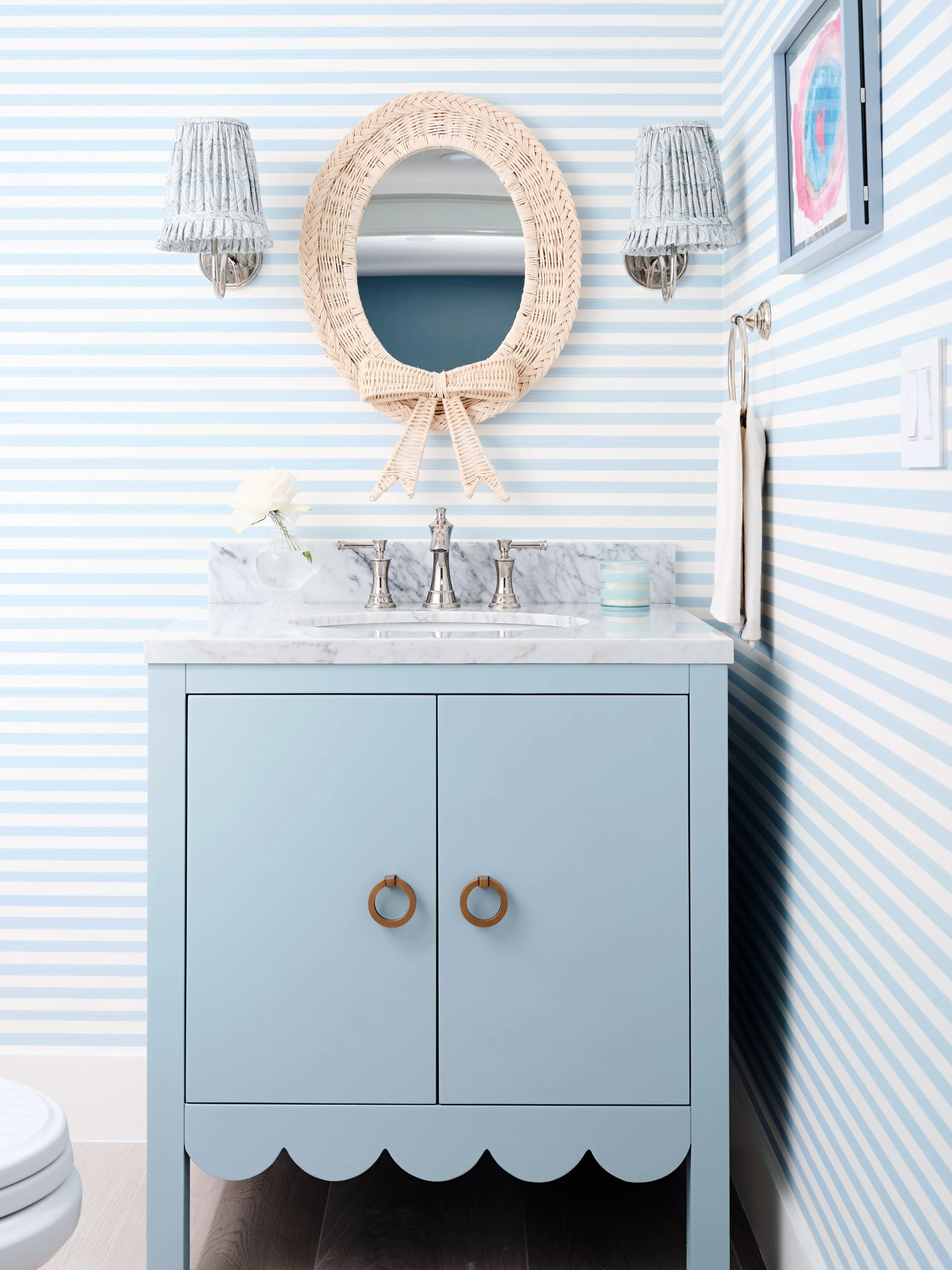 Powder room perfection in Palm Beach! 
We love designing powder rooms - a preppy horizontal  striped wallpaper, beautiful custom floral lampshades, a woven textured mirror and a powder blue scalloped vanity create a coastal playful Palm Beach combina