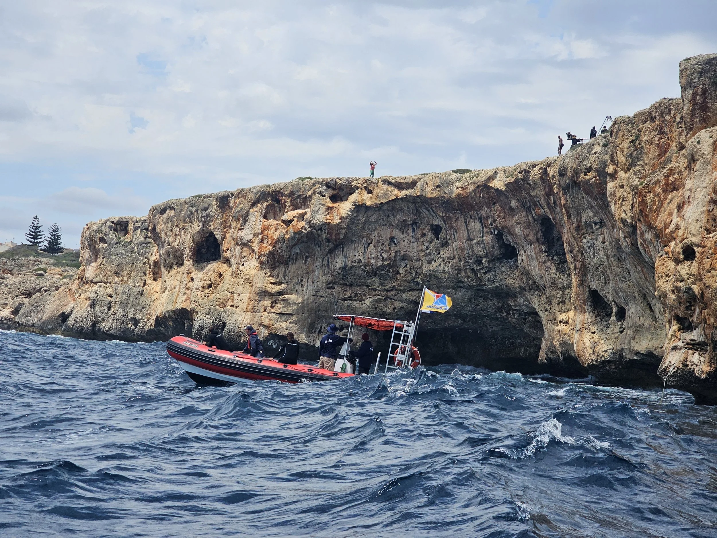 A group of people in a boat near rocky cliffs with some individuals standing on the rocks and others on a boat, with a person jumping off the rocks into the water, and film crew equipment on the rocks.