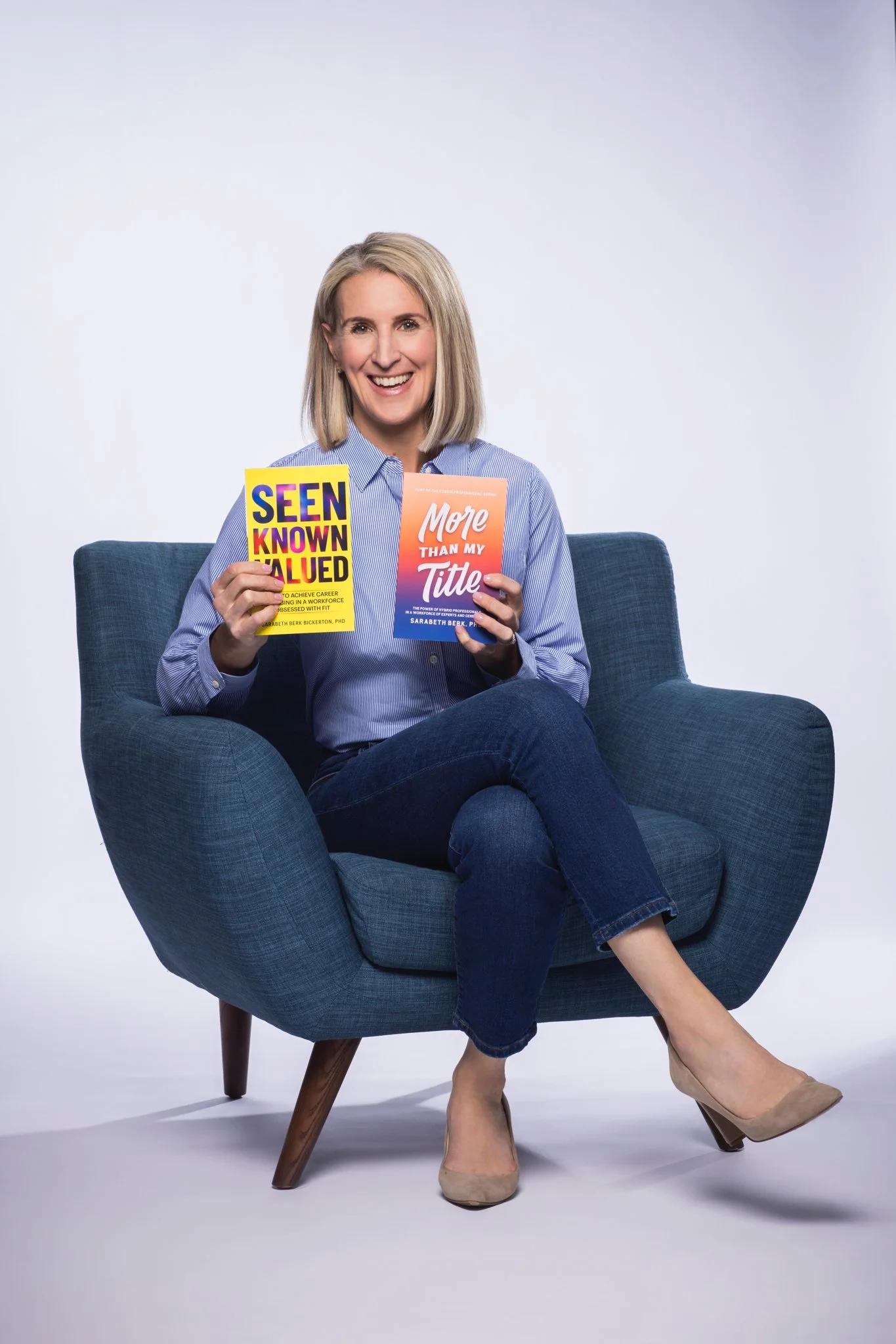 Sarabeth sits in a blue velvet chair, legs crossed holding copies of More Than My Title and Seen Known Valued