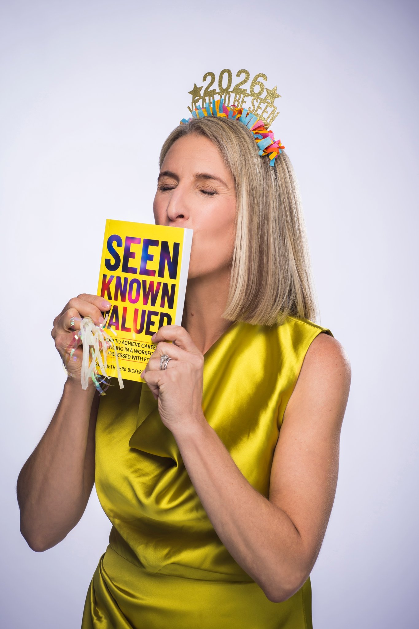 Sarabeth kisses a copy of her new book Seen Known Valued while wearing a crown and bright yellow silk dress