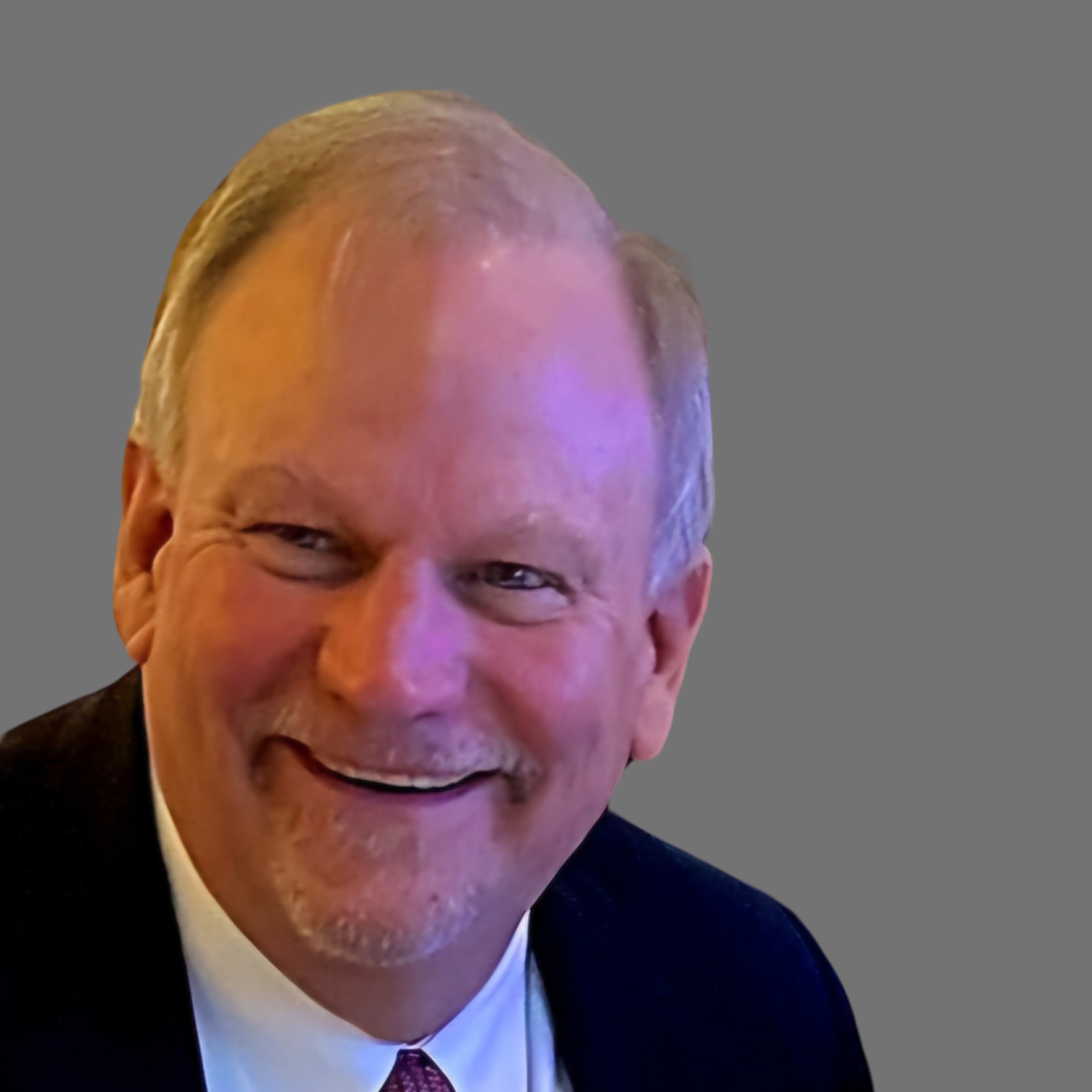 Close-up of smiling middle-aged man with light skin, gray hair, wearing a black suit, white shirt, and purple tie, against a plain gray background.