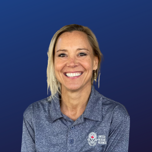 A woman with blond hair smiling, wearing a blue polo shirt with the United Rescue Alliance logo, against a dark blue background.