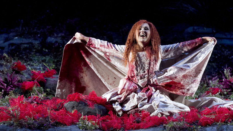 Elena Mosuc as Lucia at Teatro Regio (Turin), directed by Damiano Michieletto. Lucia kneels on the ground in a bloody wedding gown. She is surrounded by red flowers. 