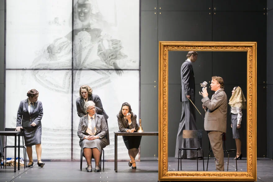 On the right, the Dwarf, the chamberlain (on stilts) and the Infanta stand inside a gold picture frame. To the left, four women in grey suits look on. Der Zwerg , Oper Graz. Photograph by Werner Kmetitsch (2016).