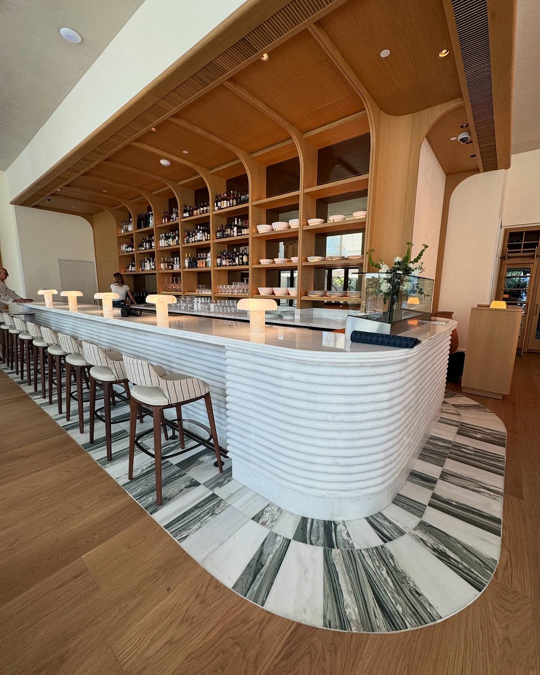 Modern bar with white textured curved counter, wooden stools with white cushions, and a back shelf with bottles and glasses, decorated with a clear glass partition and a flower arrangement.
