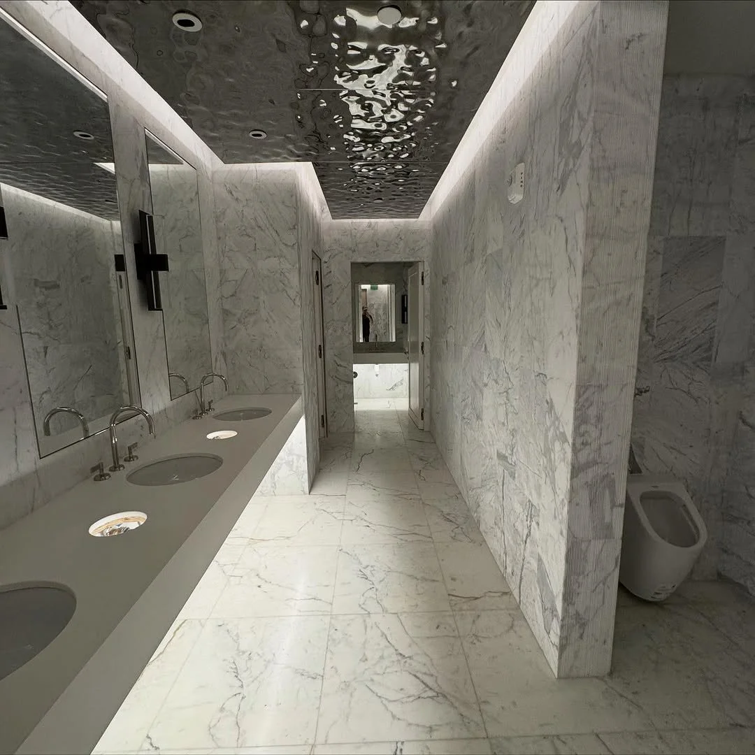 A modern bar with a marble countertop, gold-framed bar stools with tropical leaf-patterned cushions, and an array of bottles on shelves overhead, illuminated by stylish chandelier lights.