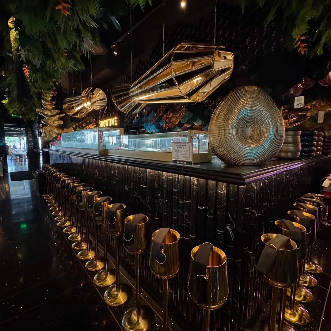 Empty upscale bar with black marble counter, gold barstools, geometric gold hanging fixtures, and decorative wall art in a dimly lit interior.