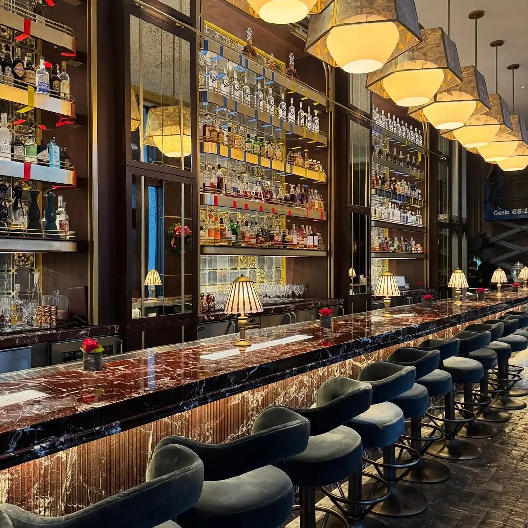 Elegant bar with marble countertop, dark wood shelves filled with liquor bottles, small lamps, and plush bar stools.