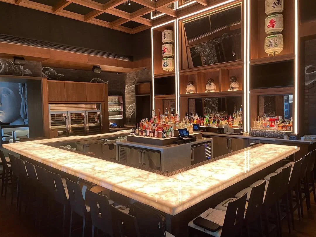 A modern restaurant bar with a glowing onyx countertop, seating around the corner, and liquor bottles on the back bar.