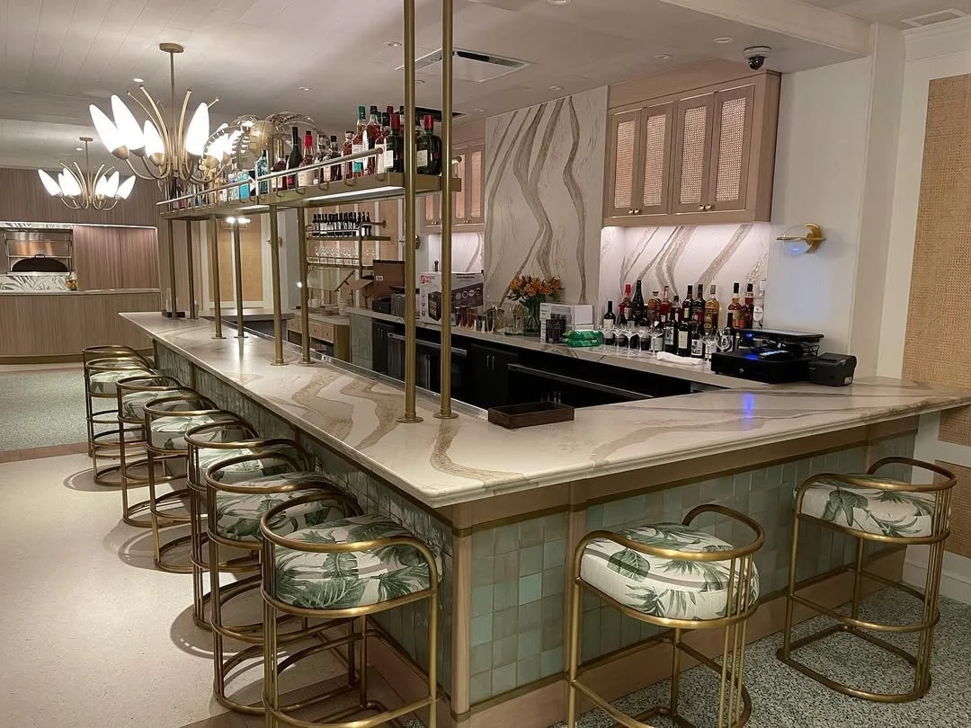 A modern bar with a marble countertop, gold-framed bar stools with tropical leaf-patterned cushions, and an array of bottles on shelves overhead, illuminated by stylish chandelier lights.