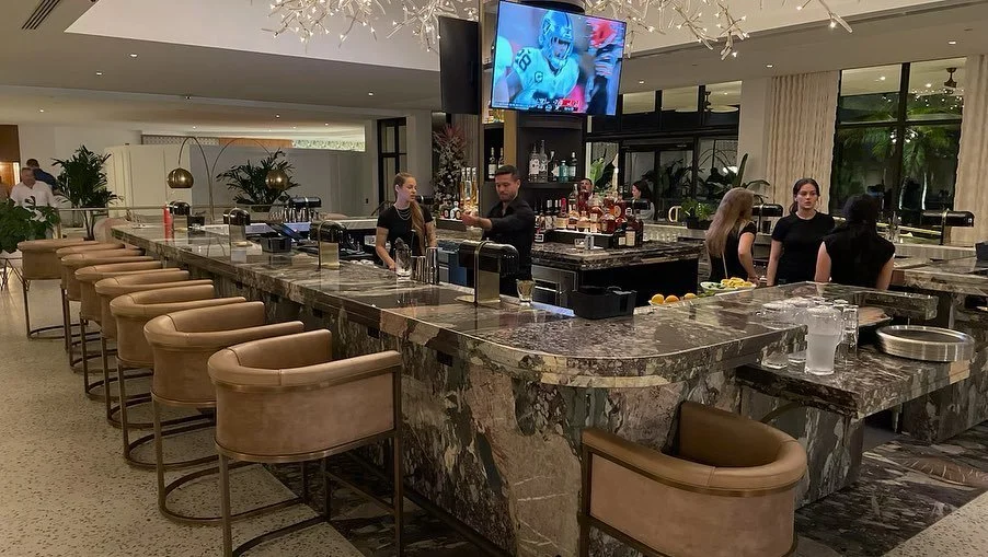 Bar with a marble countertop, high chairs, bartenders, and patrons, with a television screen showing a football game, in a modern restaurant or lounge.