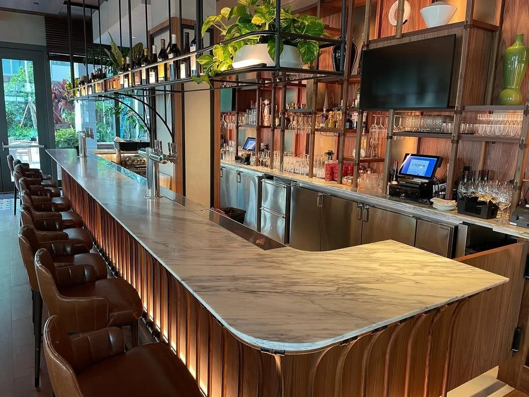 An empty bar with a marble countertop, leather chairs, wooden shelves with glassware, bottles, and bottles of wine, and a large TV screen.