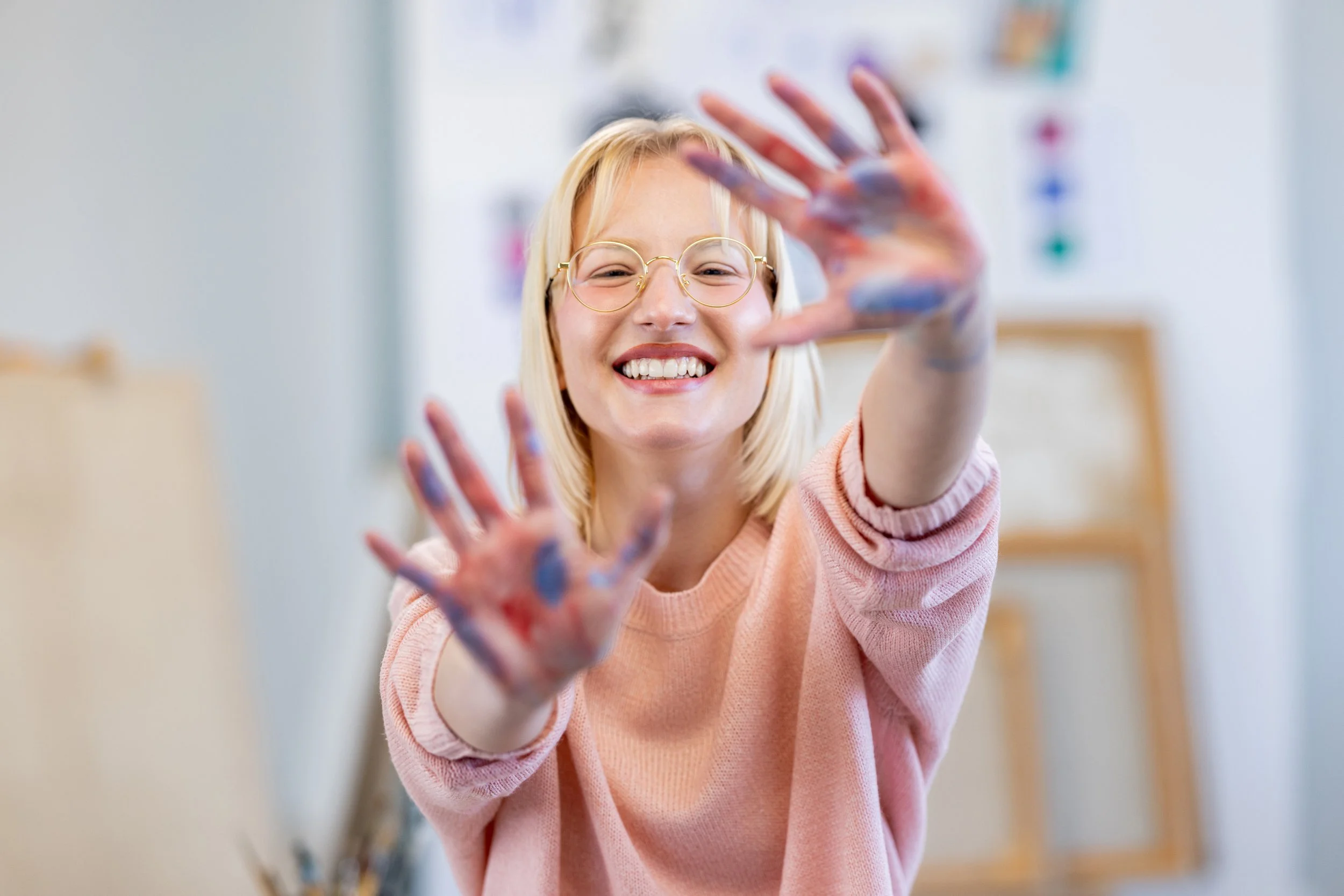 A smiling young girl with blonde hair and glasses is reaching towards the camera with her hands covered in colorful paint, in a bright room.
