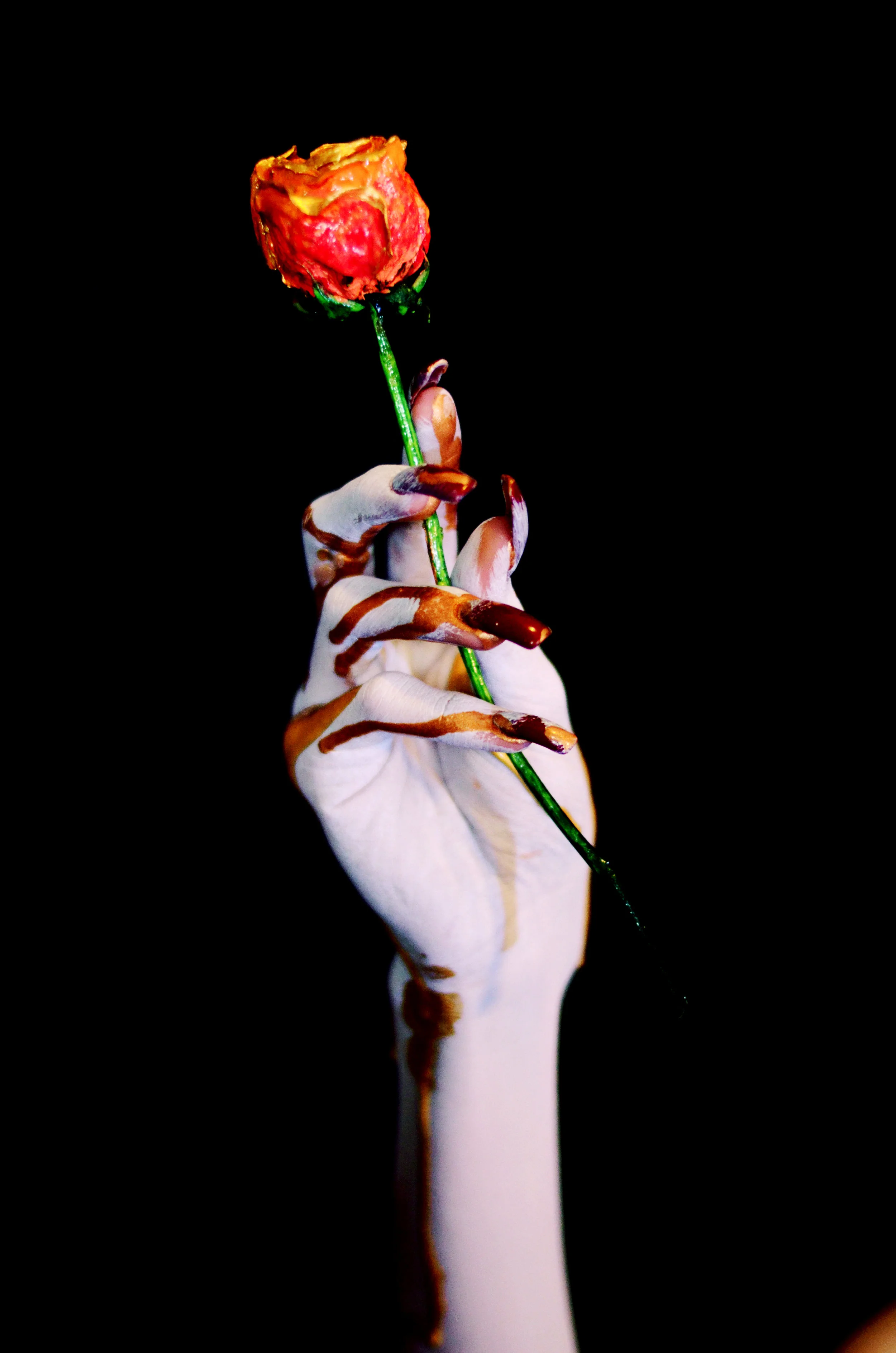 Black background with a models hand painted ghostly white, with long red nails, and gold drips of paint running down their knuckles, holding a handmade sculpture of a dried rose painted orange and red with nail polish.