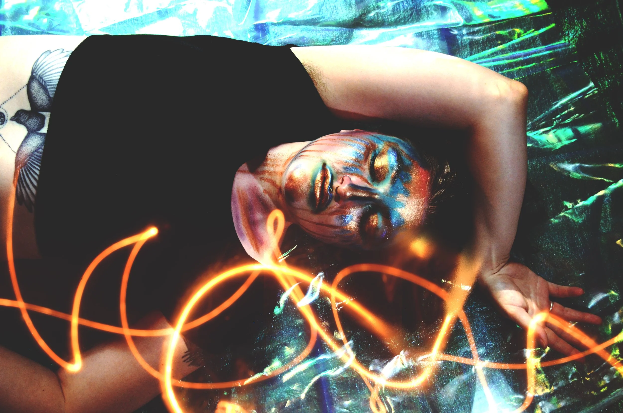 Colorful photo with a blue-green background featuring an androgynous model with copper and teal face paint and a stomach tattoo of a bird showing.