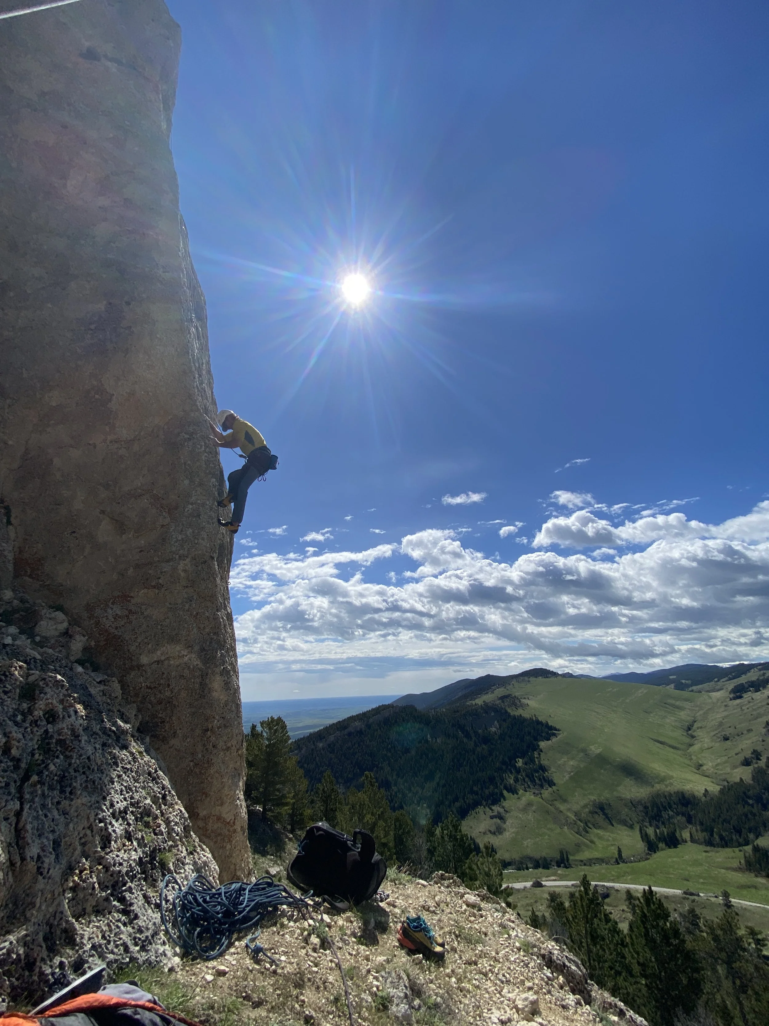 A person rock climbing on a vertical cliff under a bright sun, with a scenic view of green hills and a cloudy sky in the background.