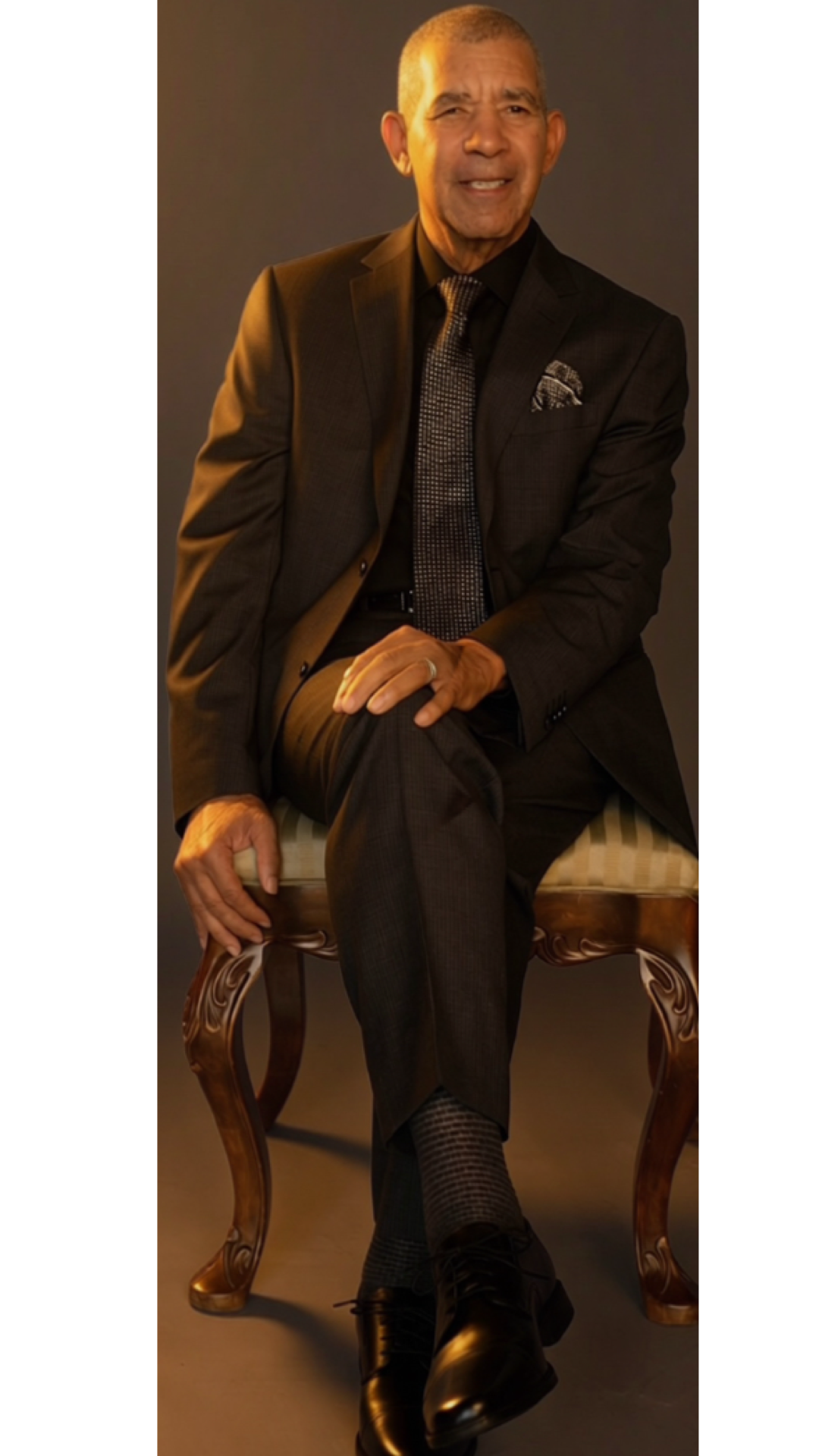 An elderly man with gray hair sitting on a wooden chair, dressed in a dark suit, patterned tie, and dress shoes, smiling at the camera against a plain background.