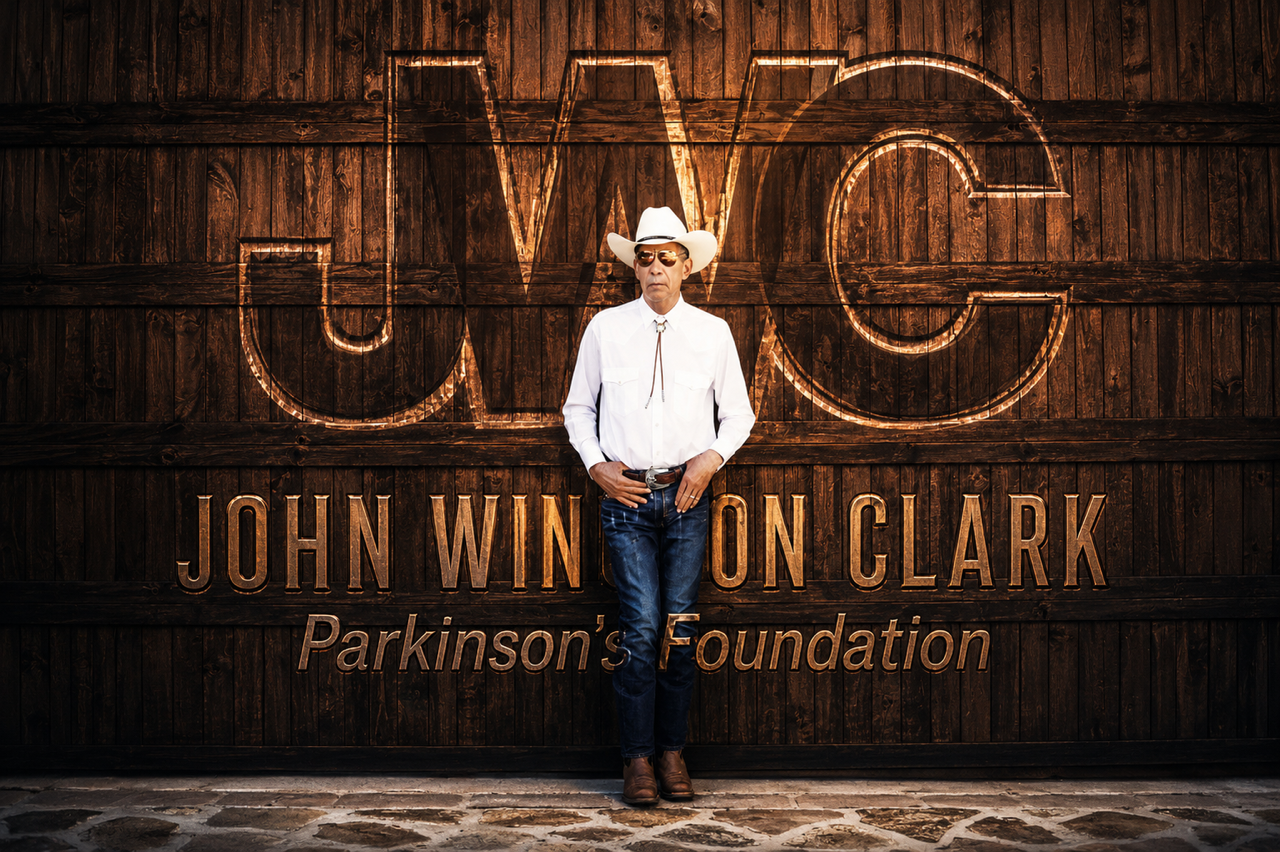 Man wearing cowboy hat, sunglasses, white shirt, and jeans standing in front of a wooden wall with large illuminated sign reading 'WC' and 'John Winon Clark Parkinson's Foundation'.