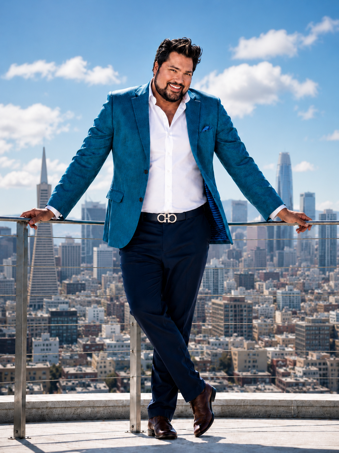 A man in a blue blazer and dark pants standing on a rooftop balcony with a cityscape background, smiling.