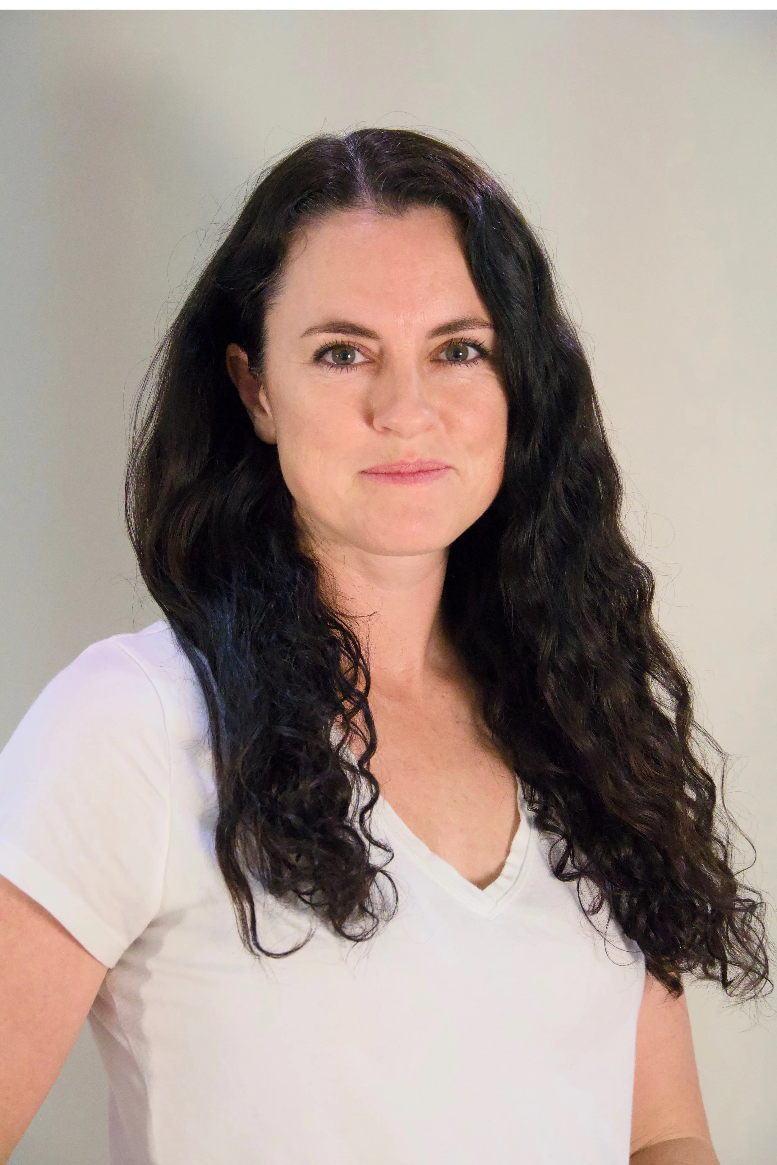 A woman with long, curly dark hair, wearing a white V-neck T-shirt, looks confidently at the camera against a plain light background.