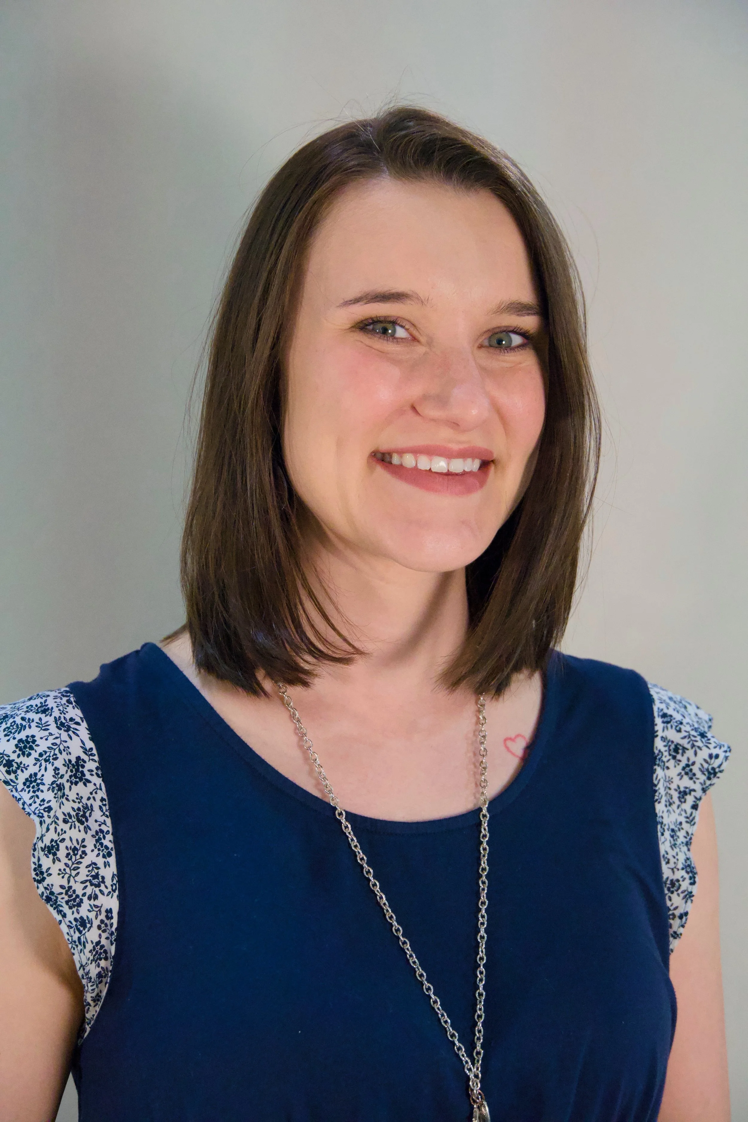 A smiling woman with shoulder-length brown hair, wearing a navy blue top with floral-patterned ruffled sleeves and a silver necklace, standing in front of a plain light gray background.