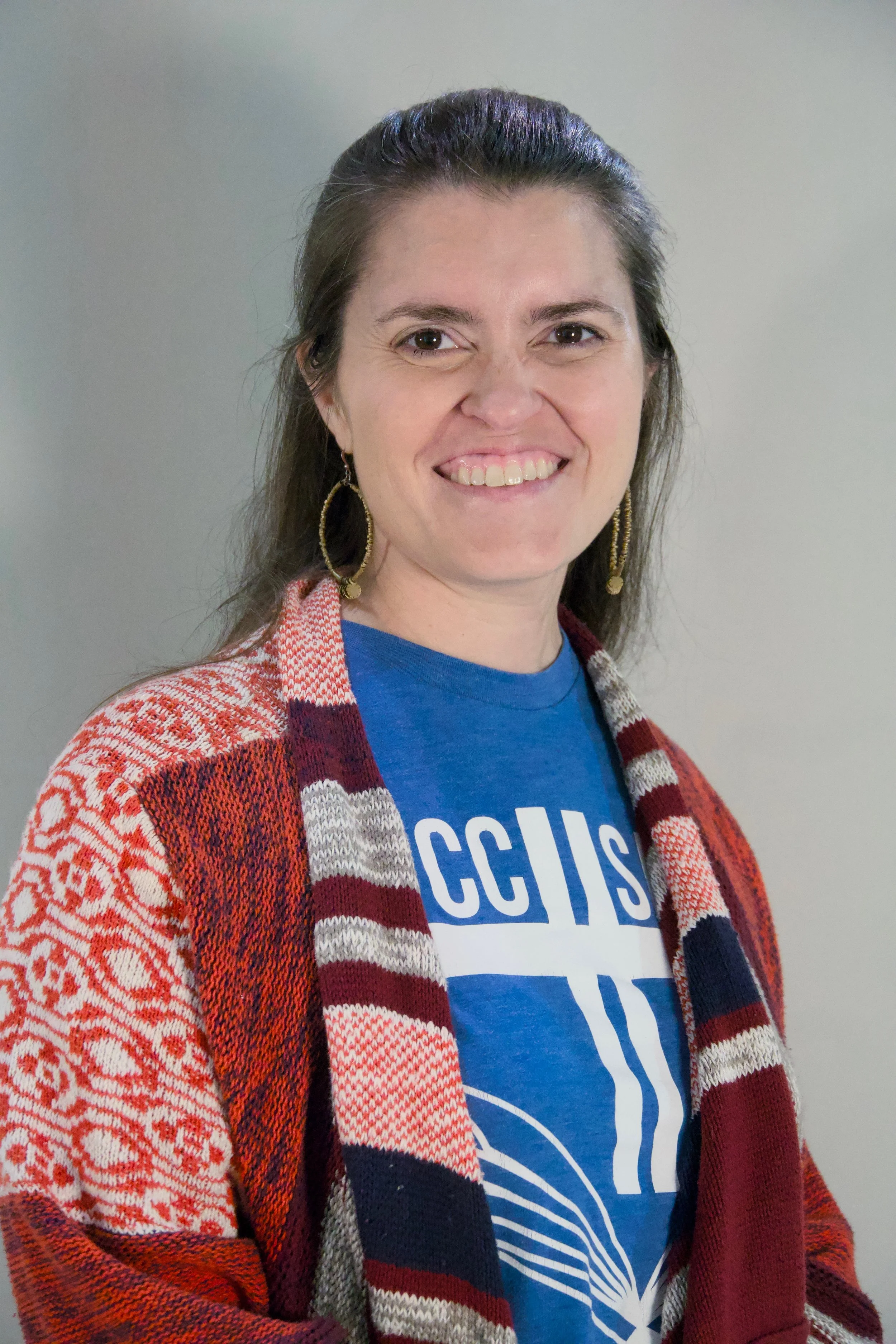 A woman with long brown hair smiling, wearing a blue t-shirt with white text and a red, white, and navy patterned cardigan.