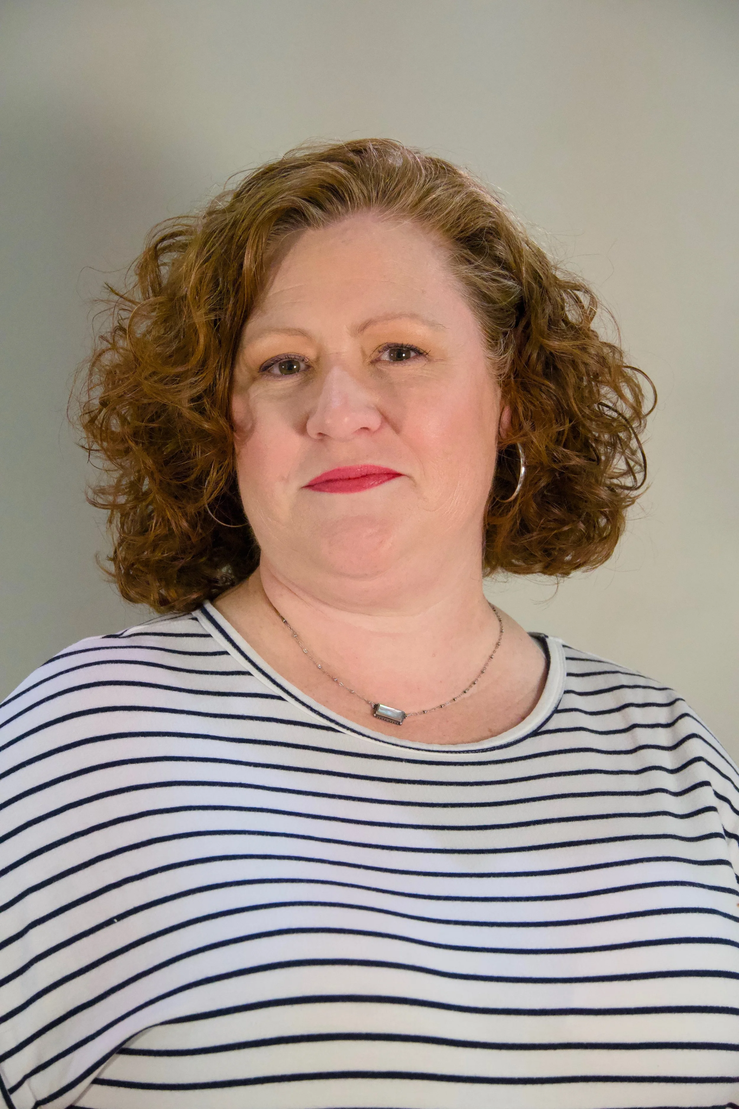 A woman with curly red hair wearing a black and white striped shirt and a necklace, standing in front of a plain wall.