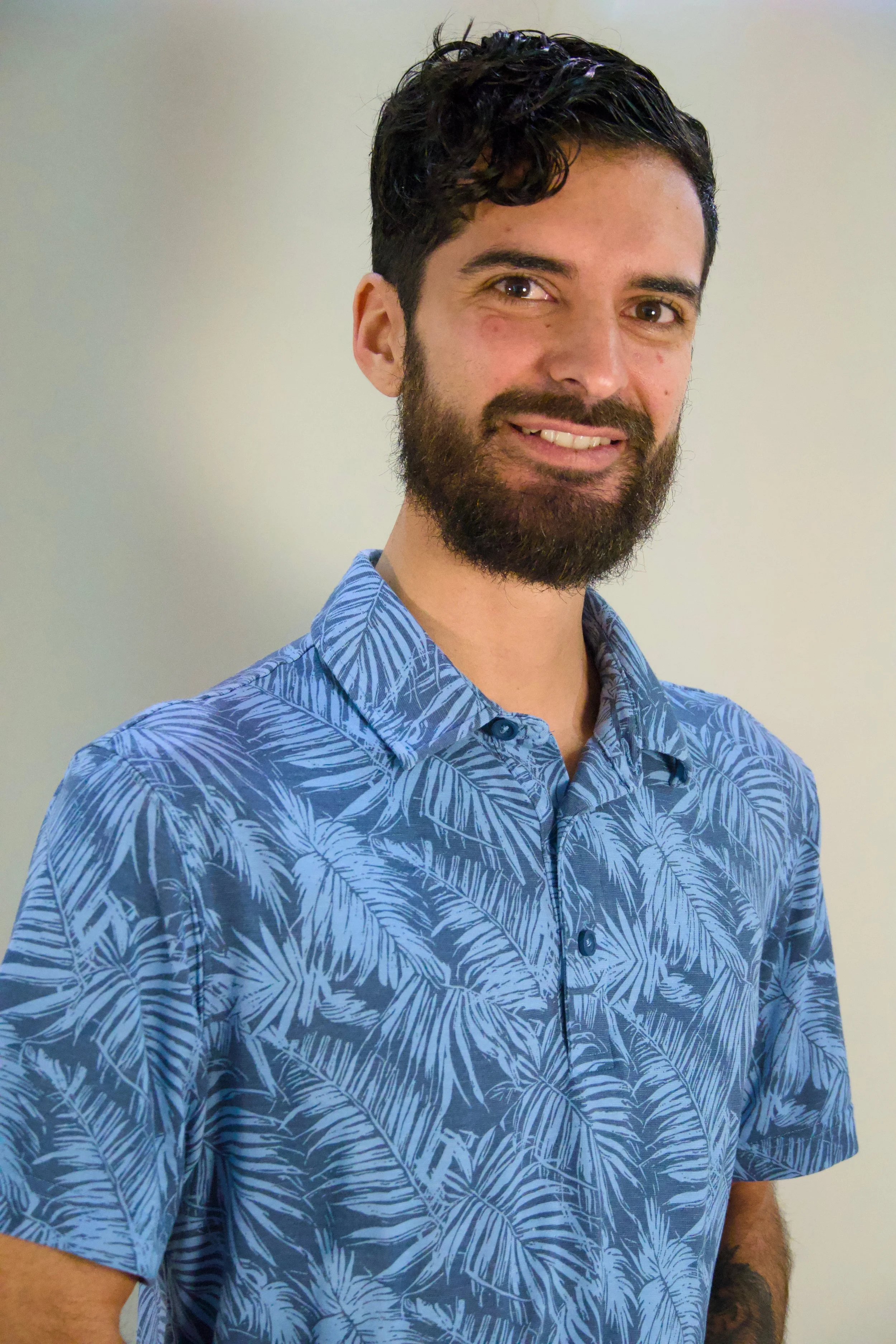 A man with dark hair and a beard smiling, wearing a blue tropical-patterned shirt, standing against a plain wall.