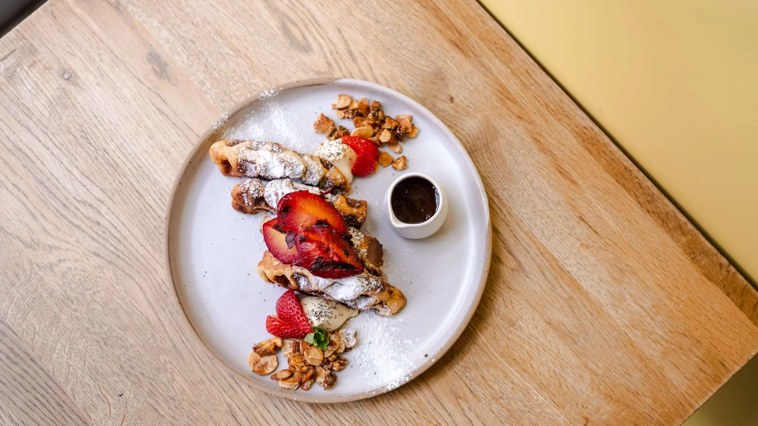 A plate of French toast topped with strawberries, powdered sugar, and syrup, served with a side of chopped nuts and sliced strawberries on a wooden table.