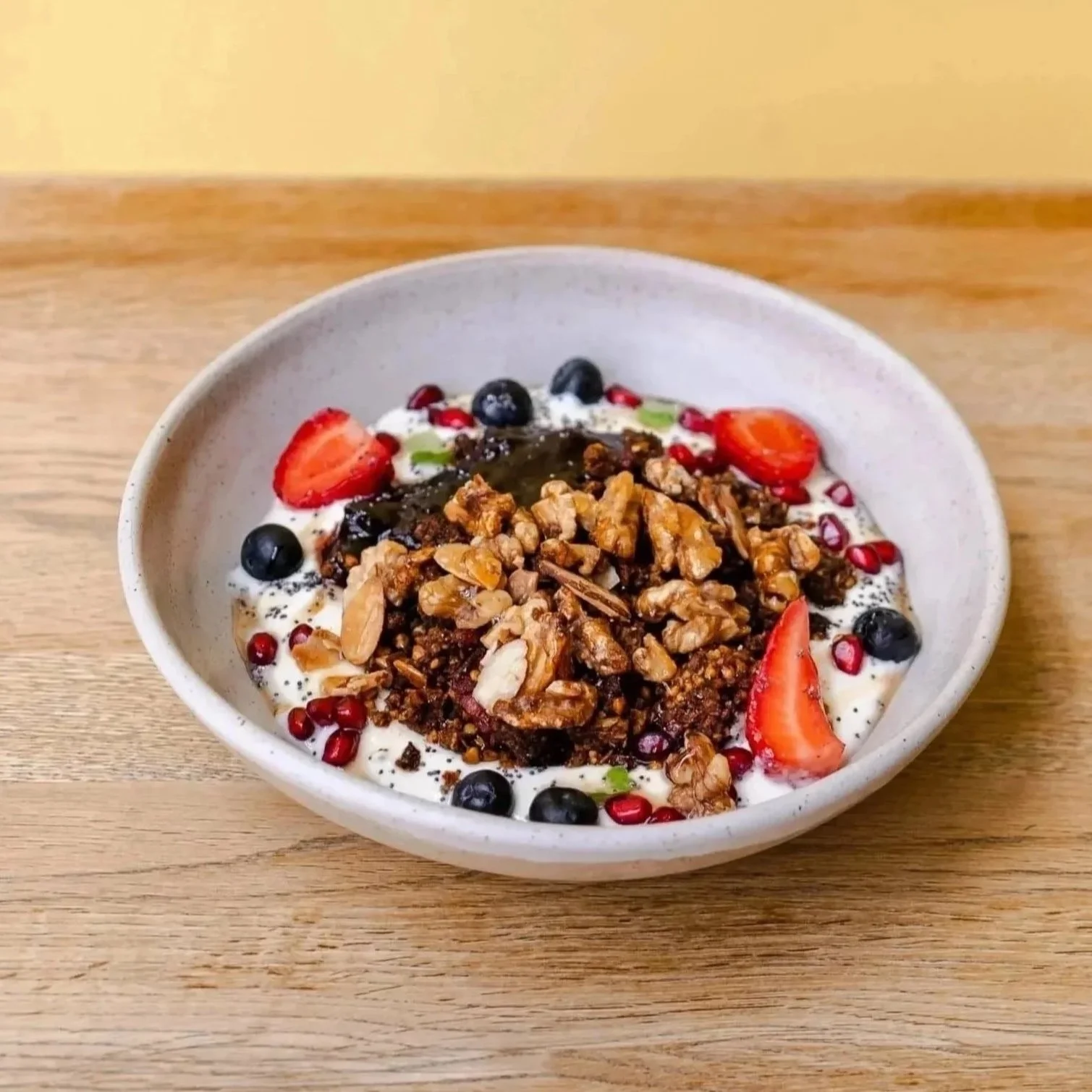 A bowl of yogurt topped with granola, sliced strawberries, blueberries, pomegranate seeds, and a drizzle of honey, placed on a wooden surface.