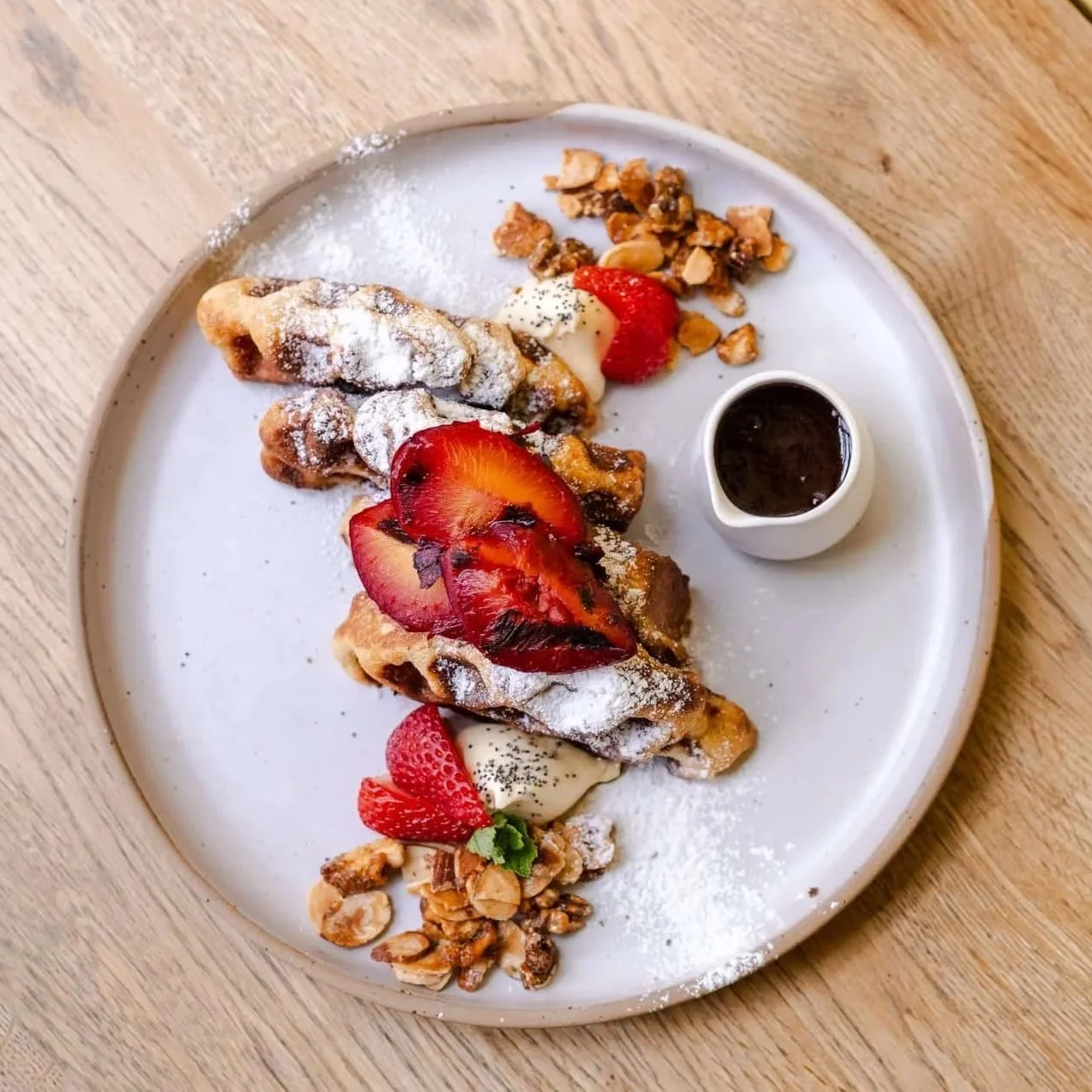A plate of French toast topped with strawberries, powdered sugar, and syrup, served with a side of chopped nuts and sliced strawberries on a wooden table.