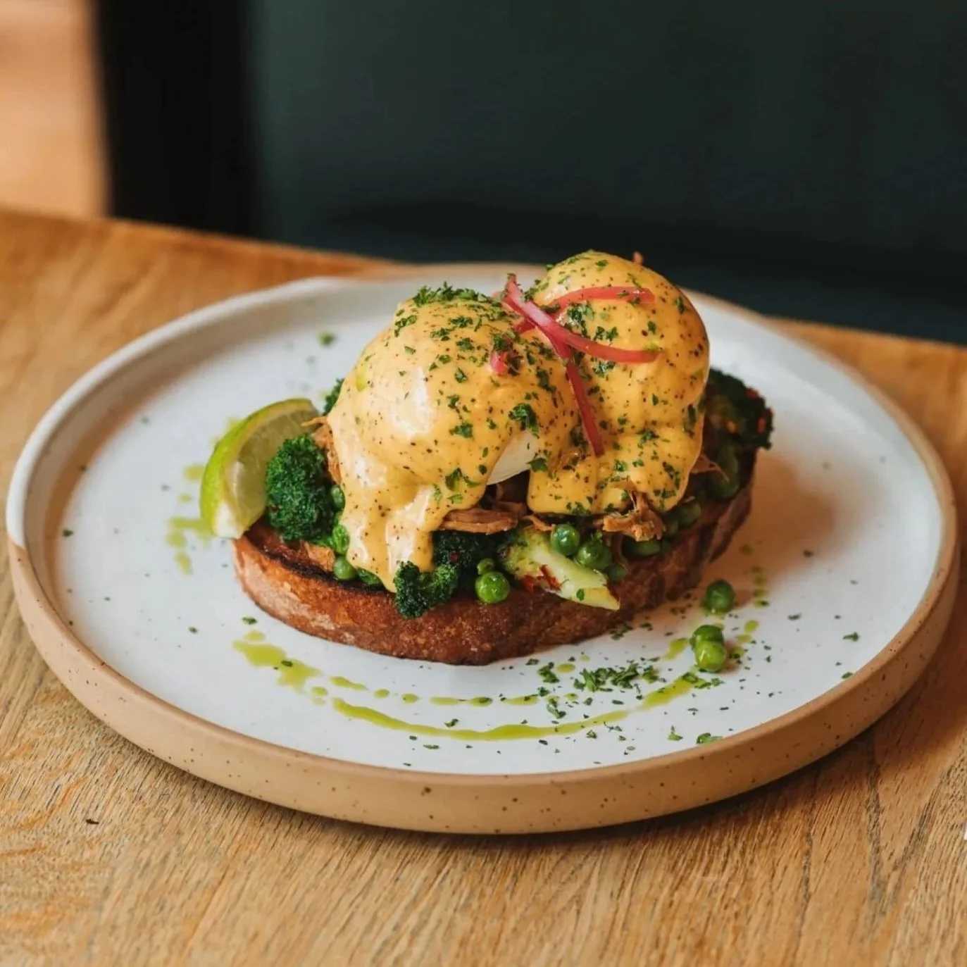 Open-faced breakfast sandwich with poached eggs, broccoli, peas, and sauce on toasted bread, garnished with herbs and a lime wedge, on a beige plate.