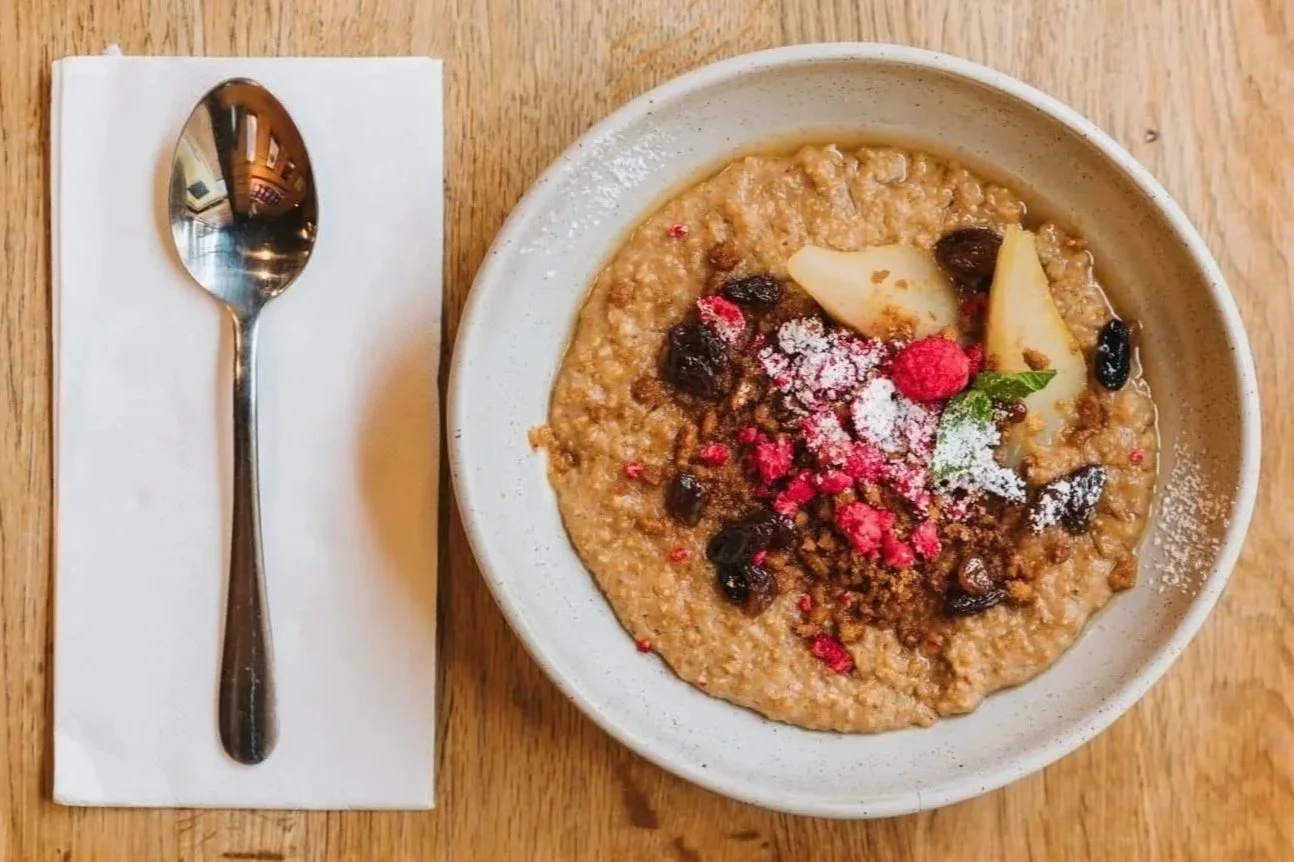 A bowl of oatmeal topped with dried berries, almond slices, powdered sugar, and pink crumb-like topping, placed on a wooden table next to a metal spoon on a napkin.