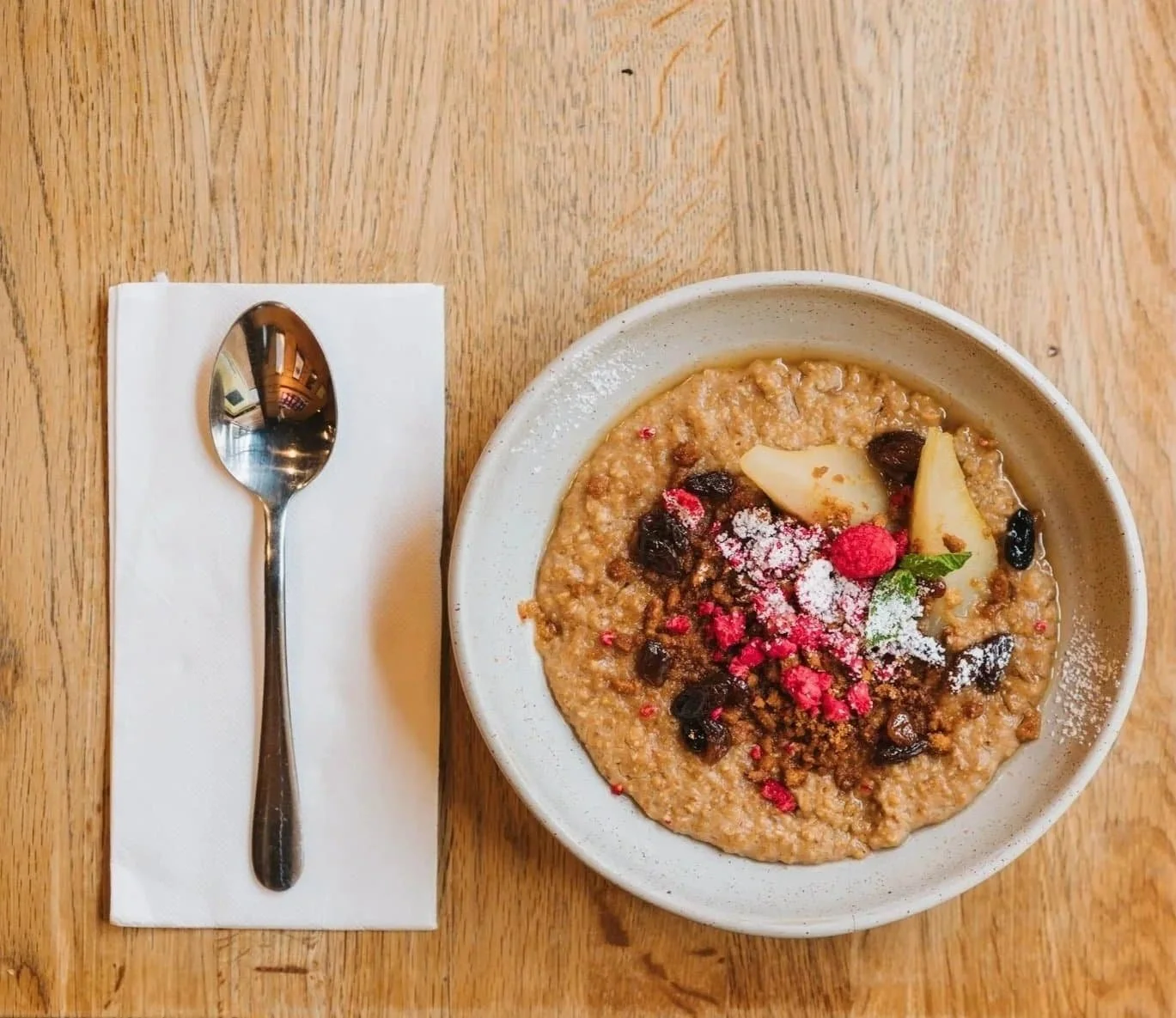 A bowl of oatmeal topped with dried berries, almond slices, powdered sugar, and pink crumb-like topping, placed on a wooden table next to a metal spoon on a napkin.
