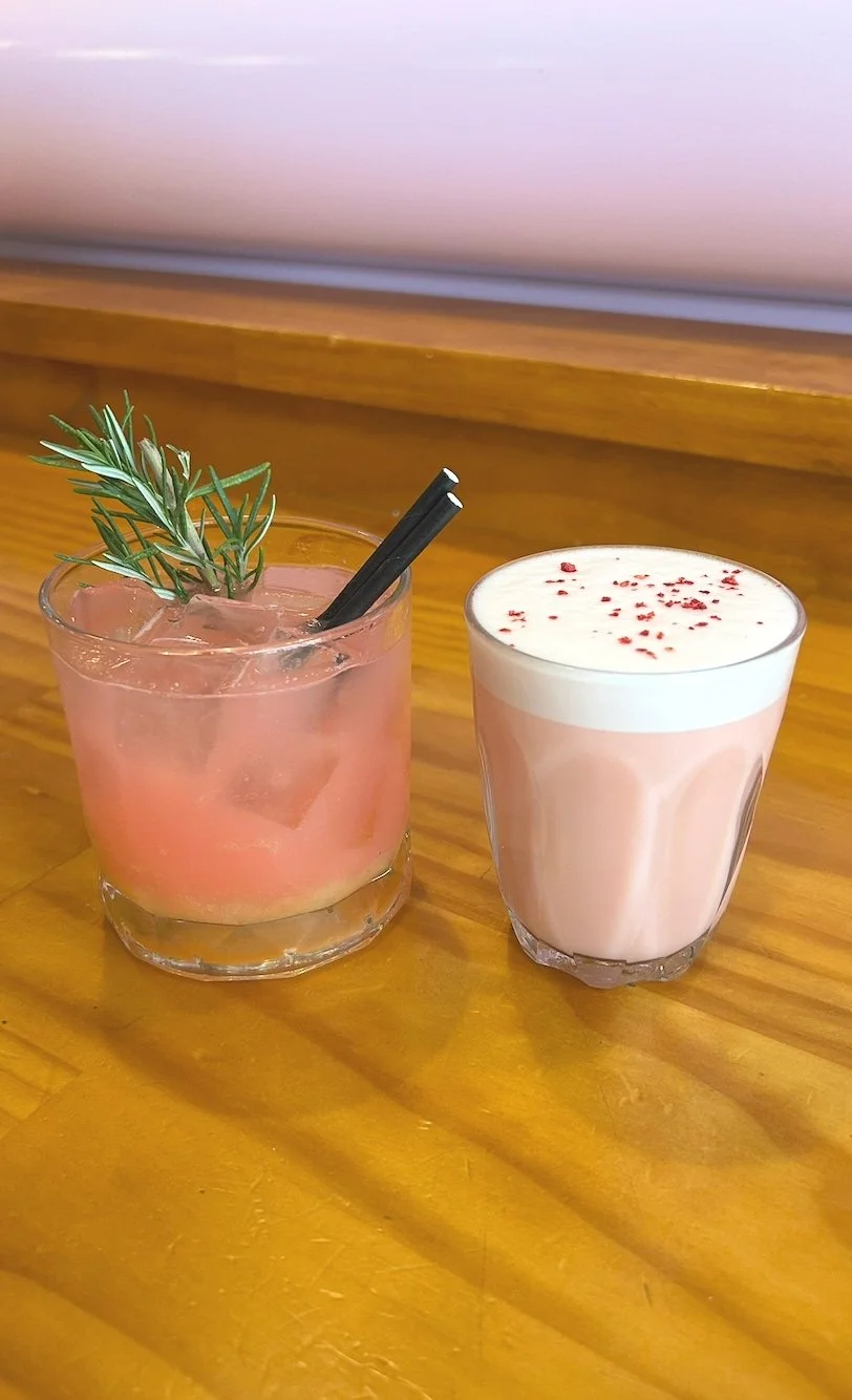 Two pink-colored beverages on a wooden table, one garnished with a sprig of rosemary and ice, the other topped with whipped cream and red sprinkles.