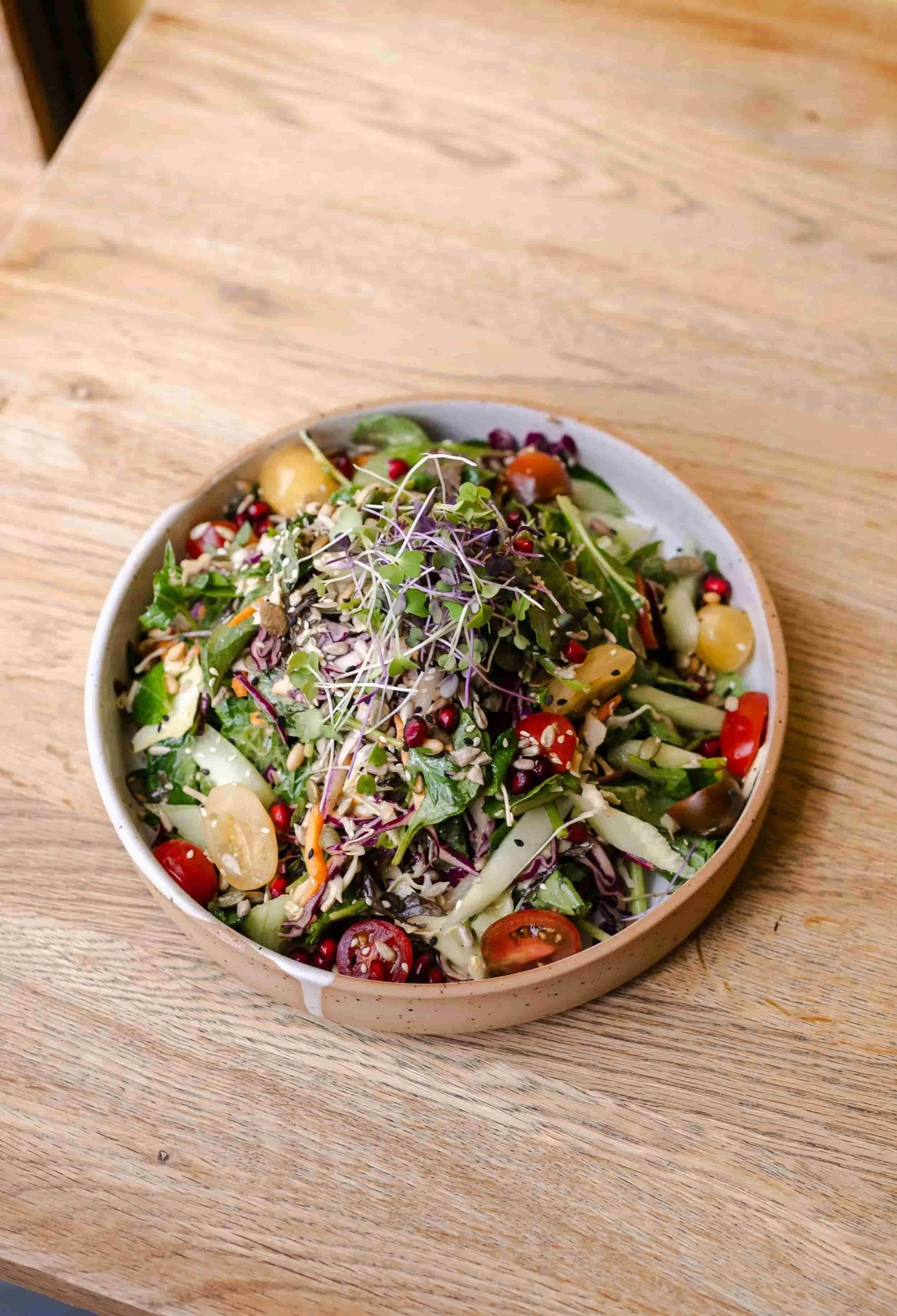 A bowl of mixed salad containing cherry tomatoes, microgreens, shredded vegetables, and sunflower seeds on a wooden table.
