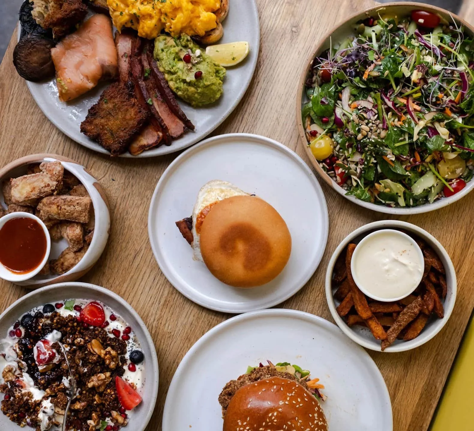 A variety of dishes on a wooden table including salad, fried chicken, burgers, roasted vegetables, hummus, and a yogurt dessert with berries.