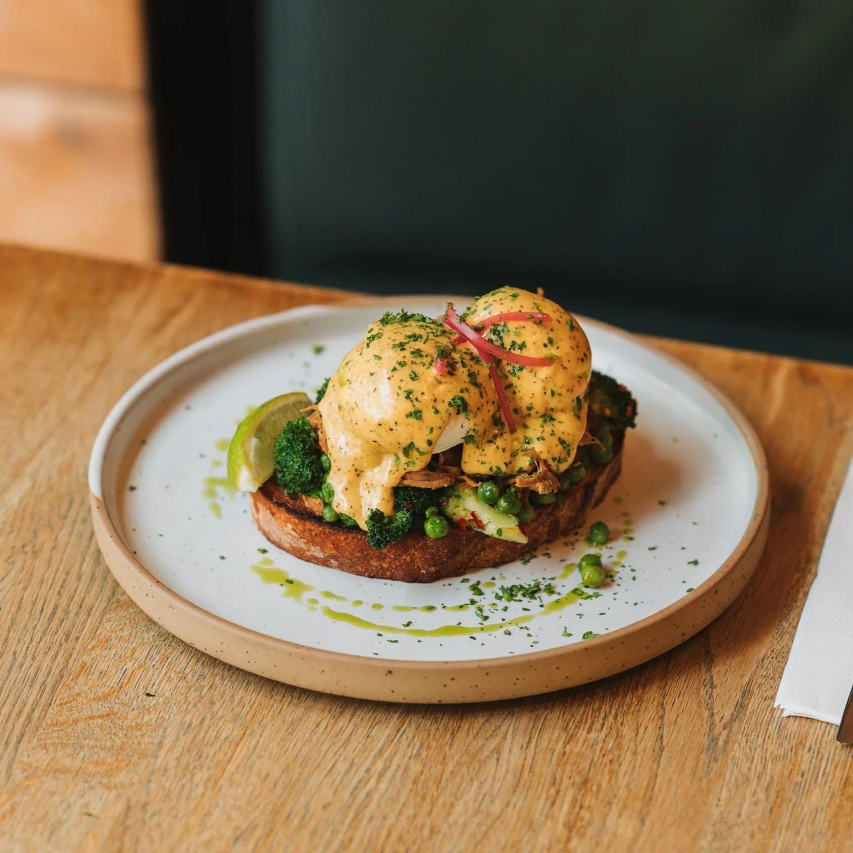 Open-faced breakfast sandwich with poached eggs, broccoli, peas, and sauce on toasted bread, garnished with herbs and a lime wedge, on a beige plate.