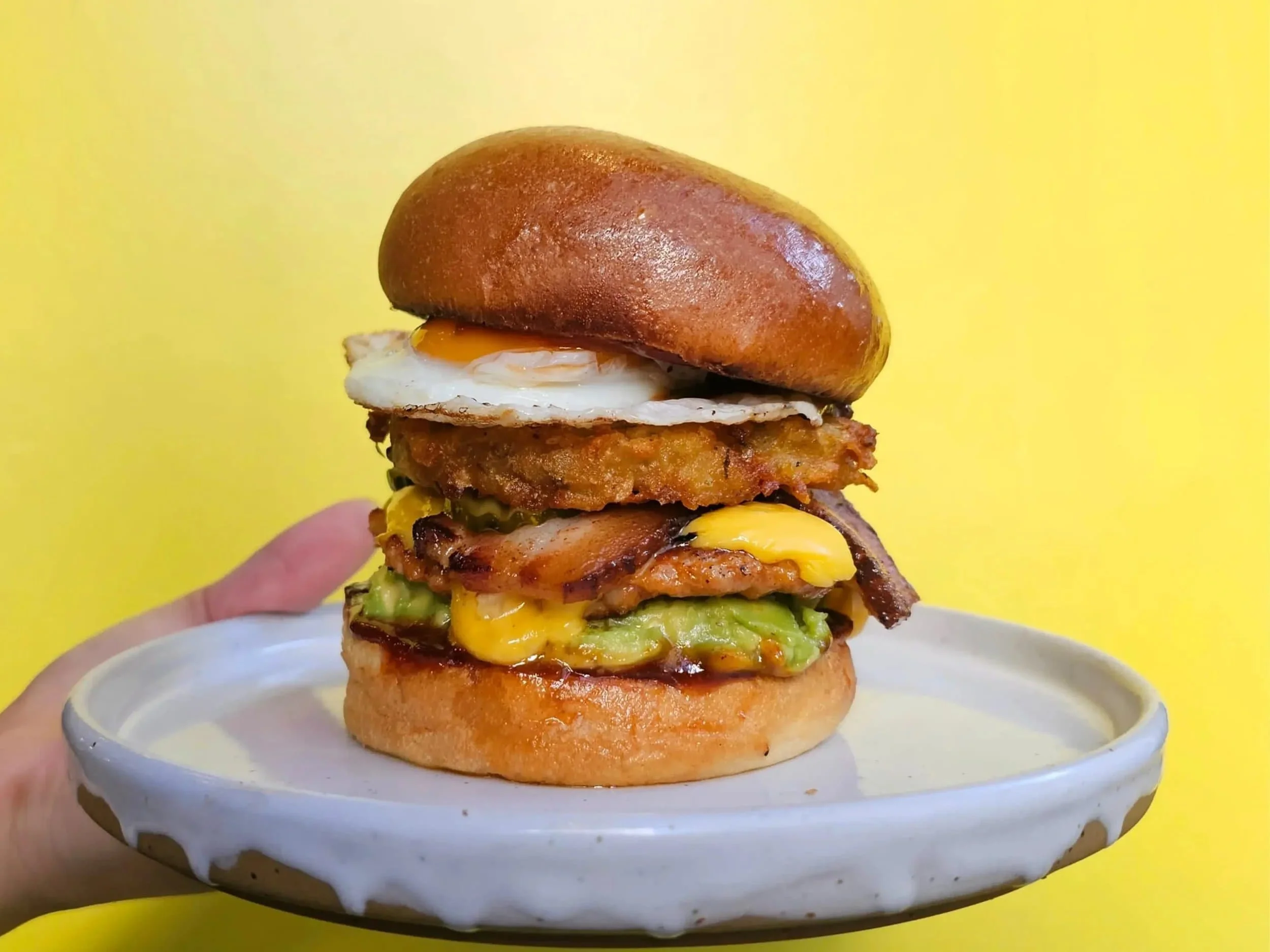 A large burger with a glossy brioche bun, topped with a fried egg, crispy chicken fillet, bacon, cheddar cheese, and avocado slices, served on a white plate against a yellow background.