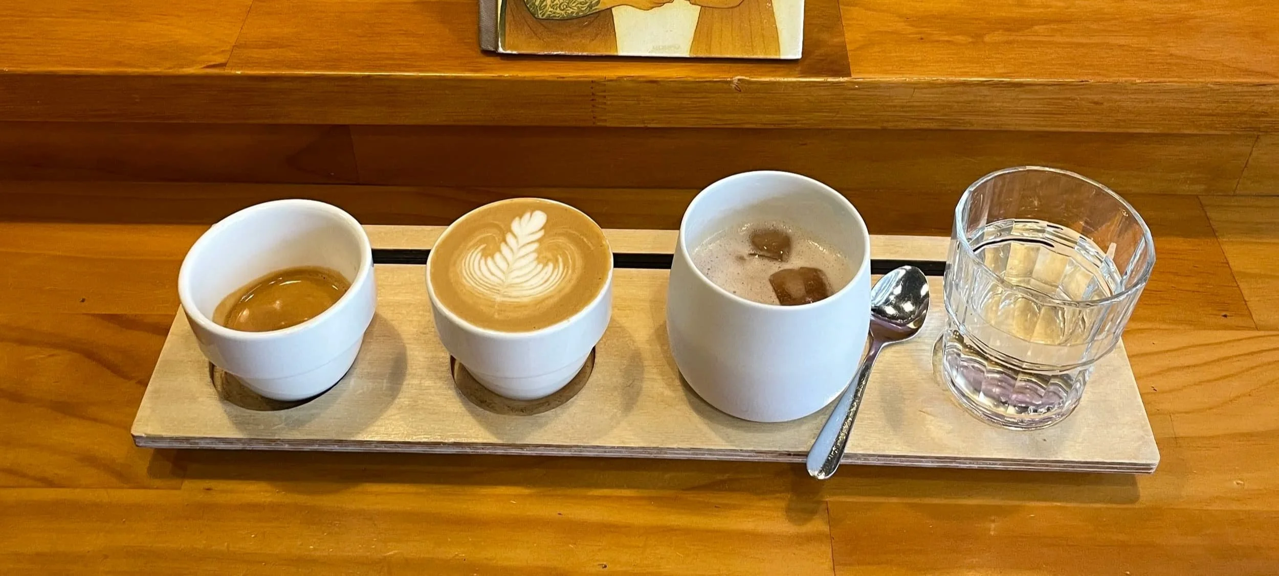 Four cups with different beverages, a glass of water, and a spoon on a wooden tray on a wooden table.