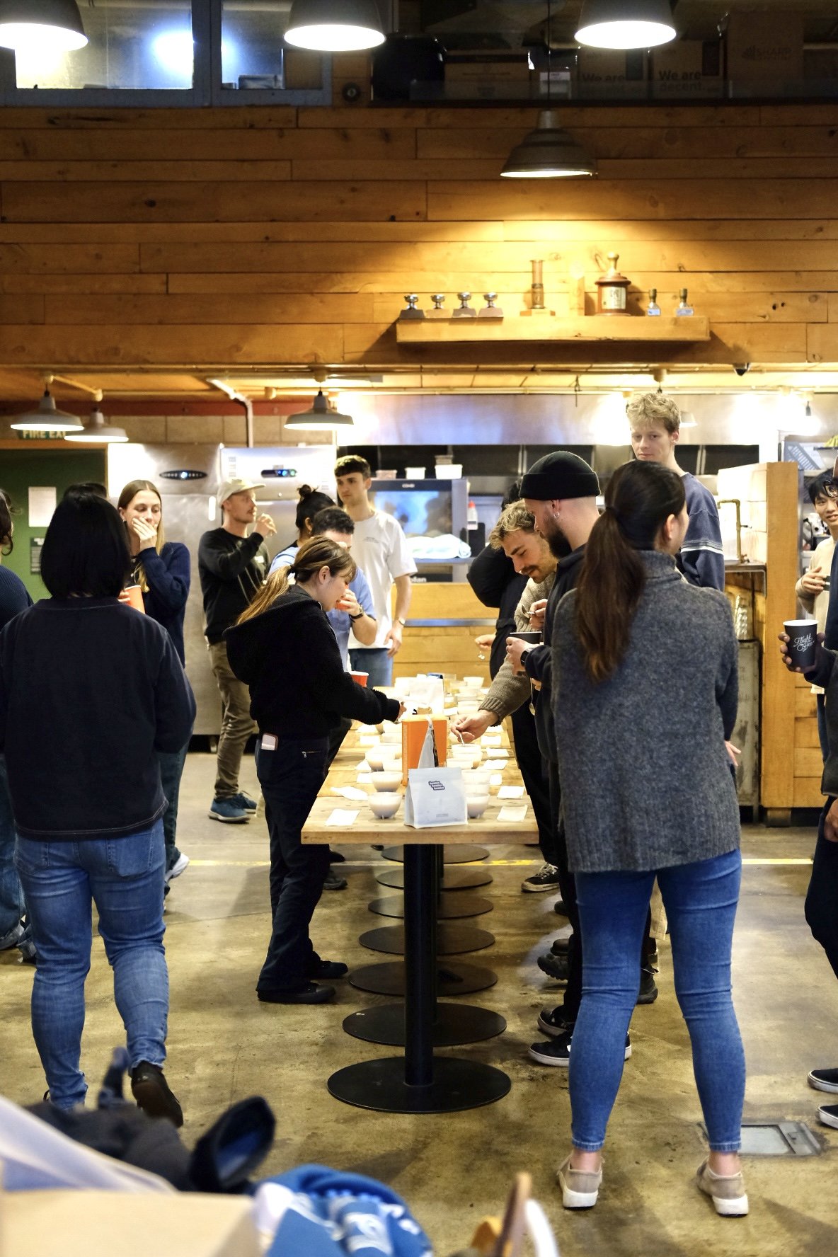 People gathered around a table with bowls and cards in a coffee shop or cafe setting, some tasting or evaluating food or drinks.